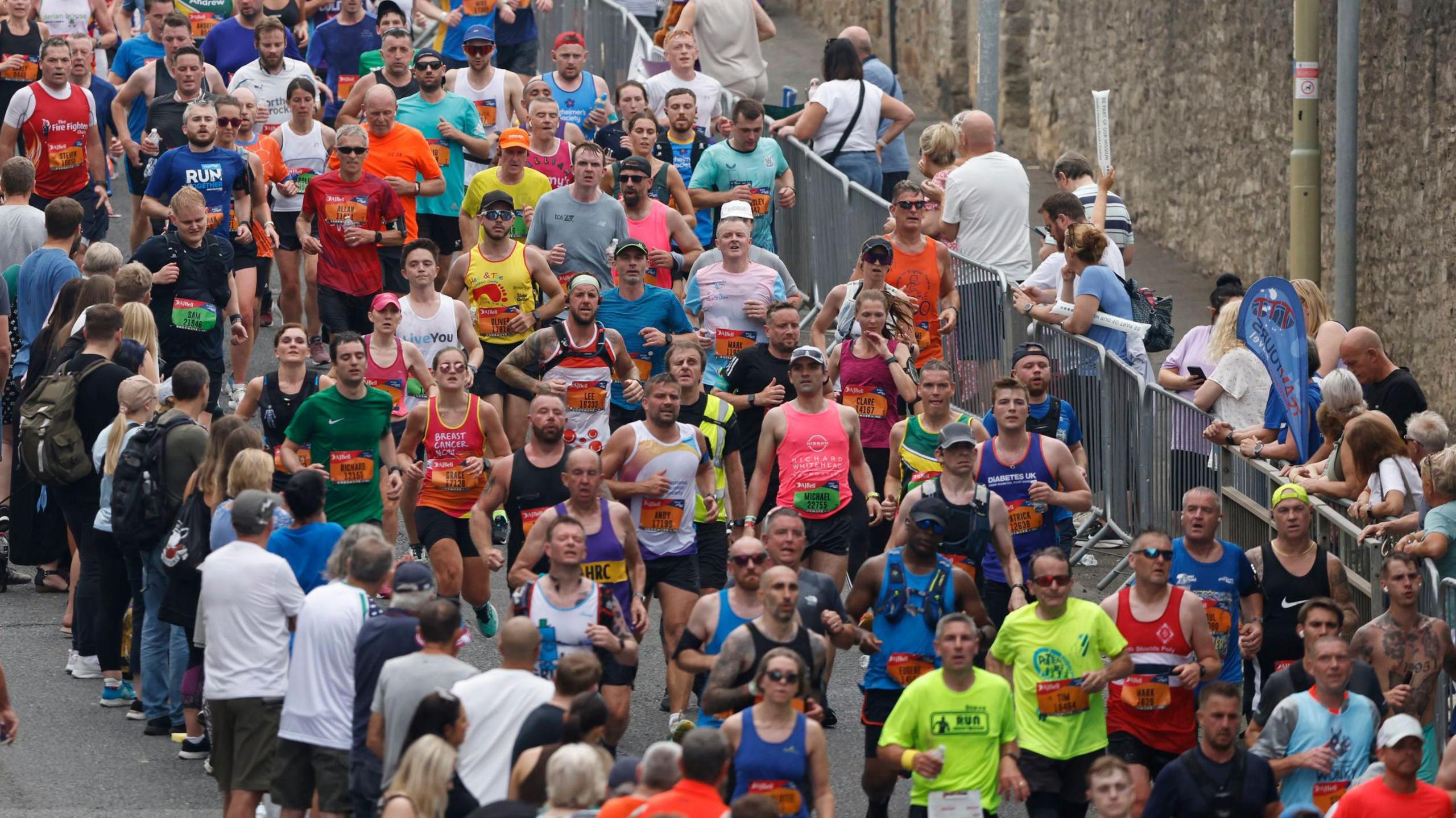 Crowds of runners, wearing colourful race vests, run down a road as crowds cheer them on.