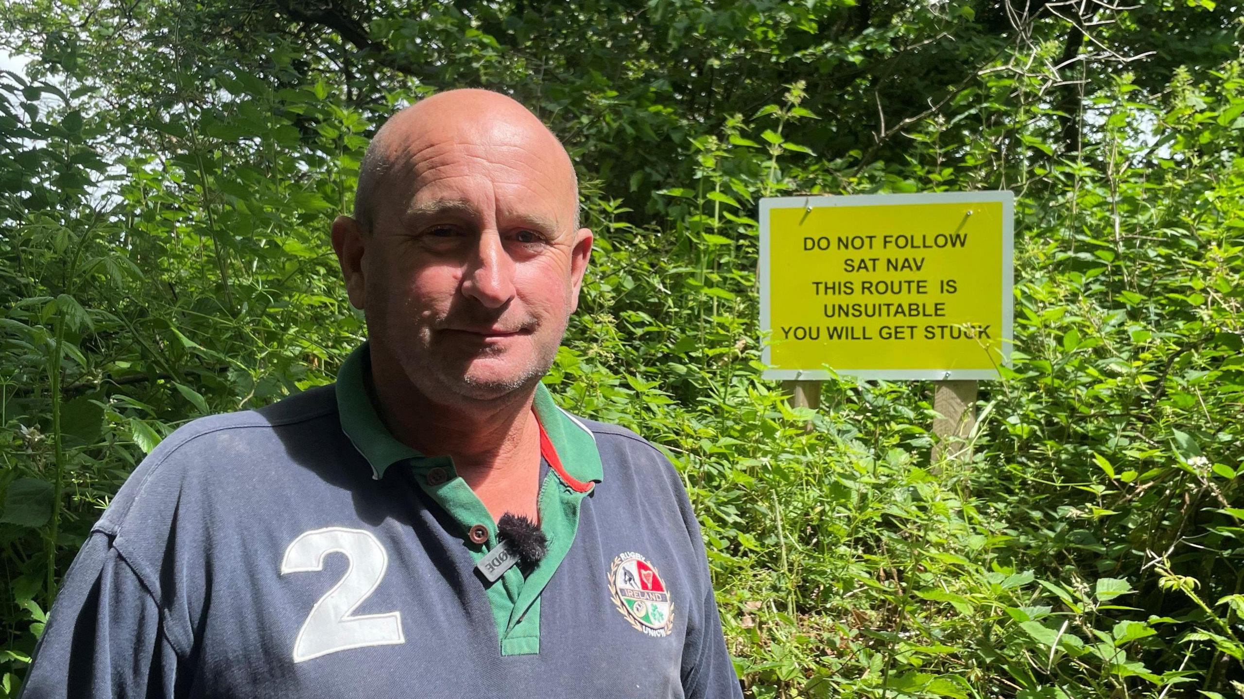 Mark Nicholls wearing a grey T-shirt stood in front of a hedge. In the hedge is one of his signs which reads: "Do not follow sat-nav this route is unsuitable you will get stuck."