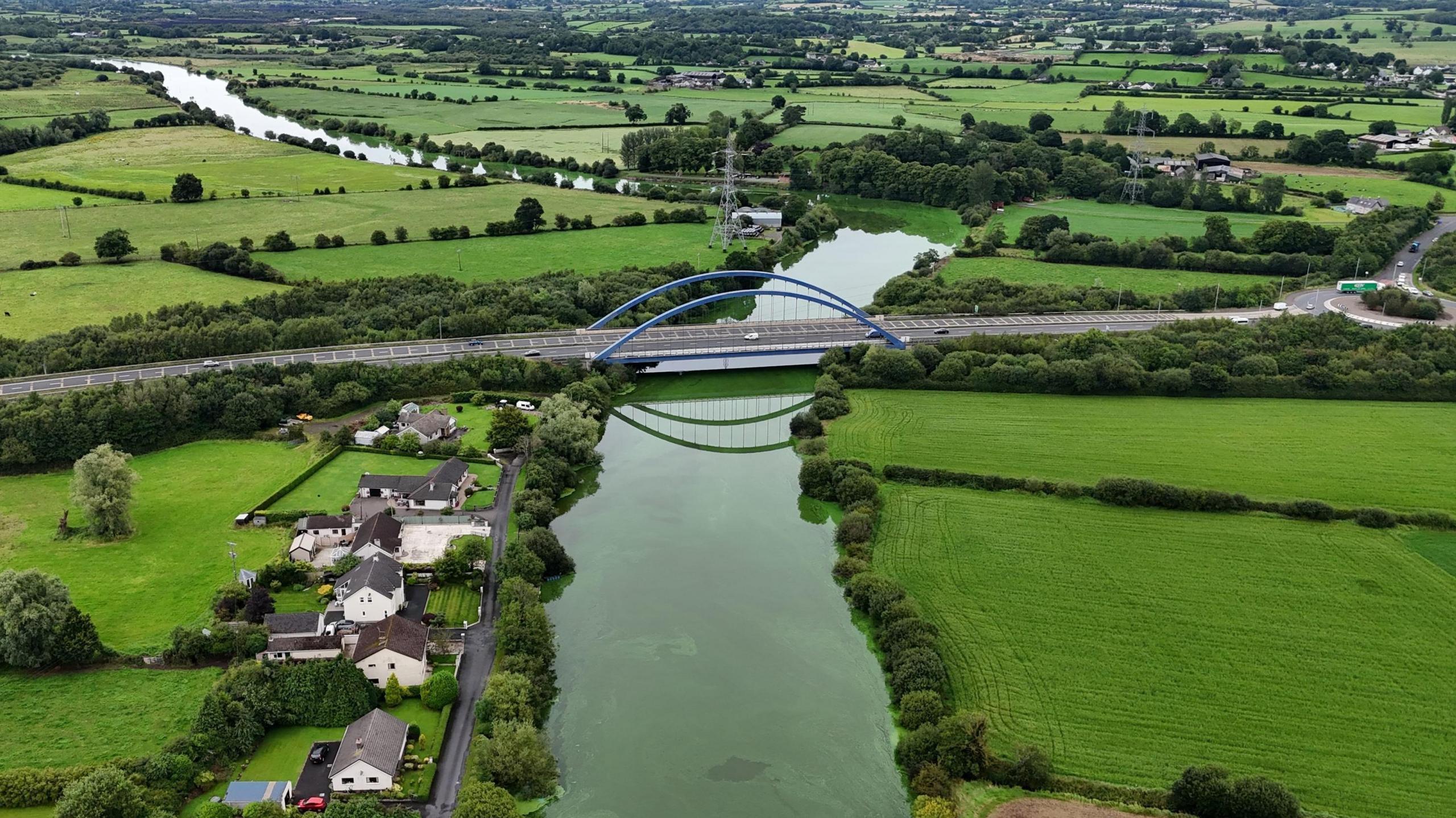 An aerial photo of a bridge across the River Bann where it meets Lough Neagh near the village of Toome.  The river appears almost as green as the surrounding fields due to blue-green algae.