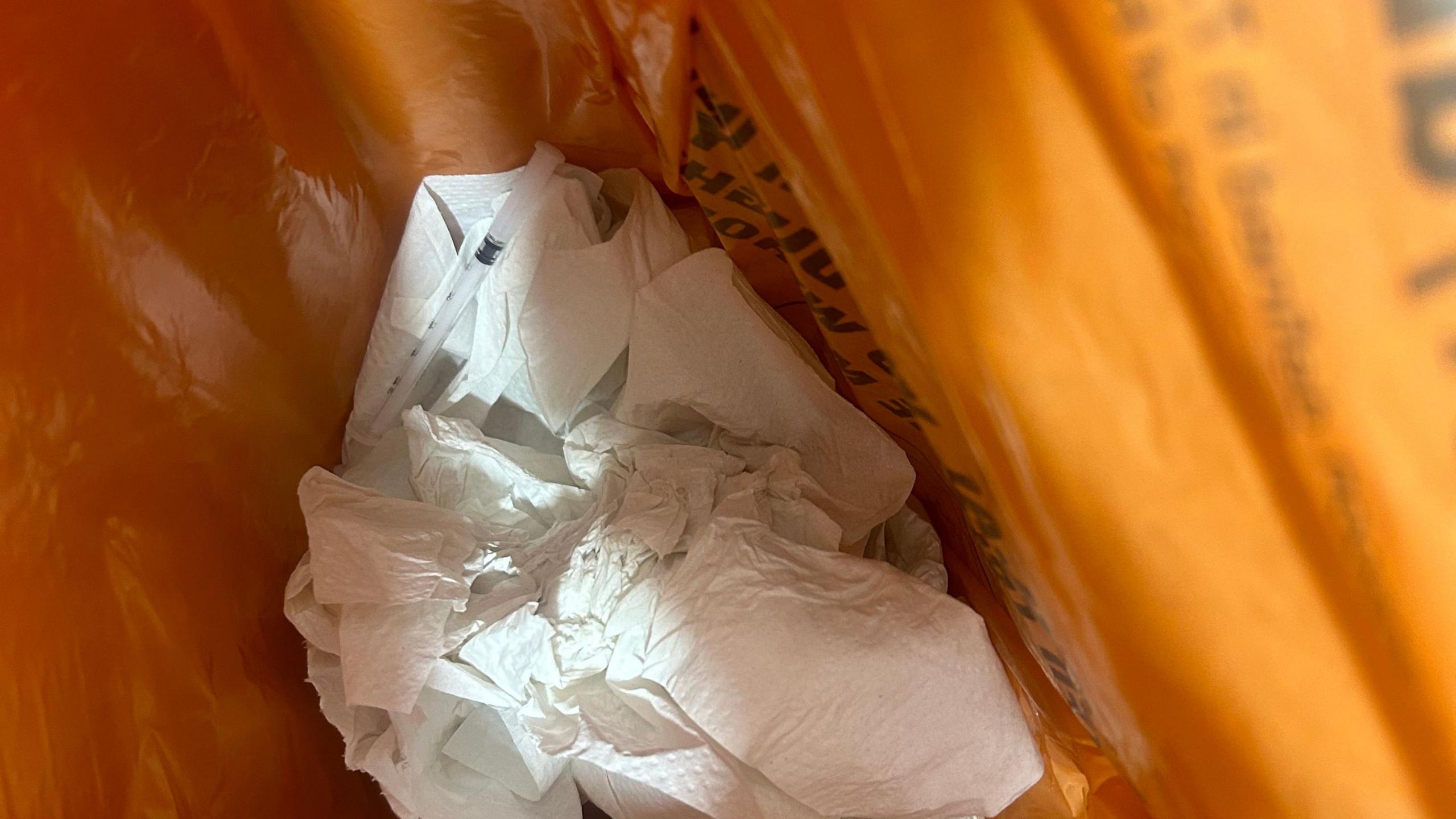 A landscape photo of a waste basket in one of the emergency department toilet bathrooms. There are multiple scrunched up pieces of tissue as well as a discarded needle.