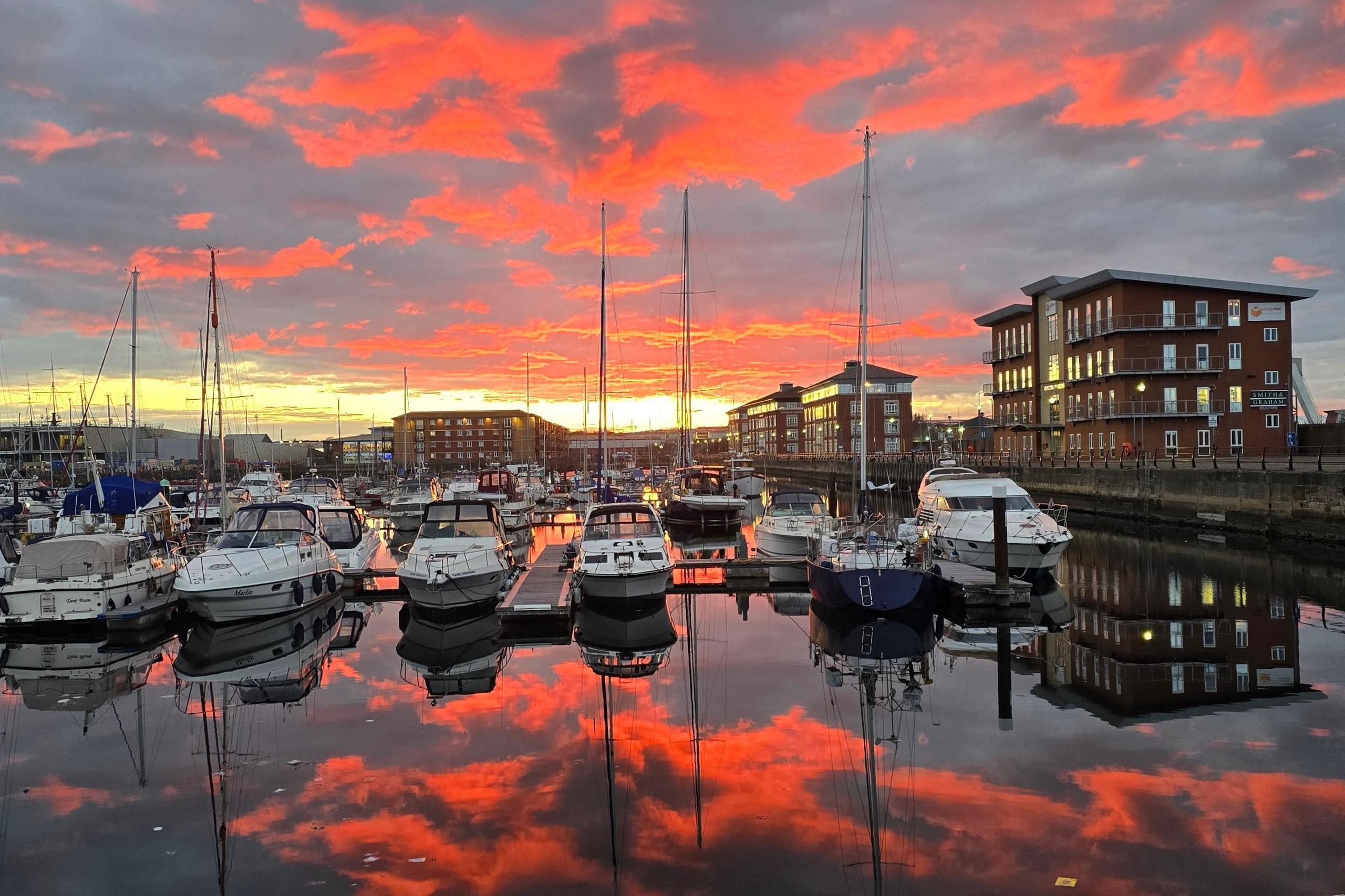 The clouds are illuminated orange at sunset over the marina. The sky is reflected back in the water.