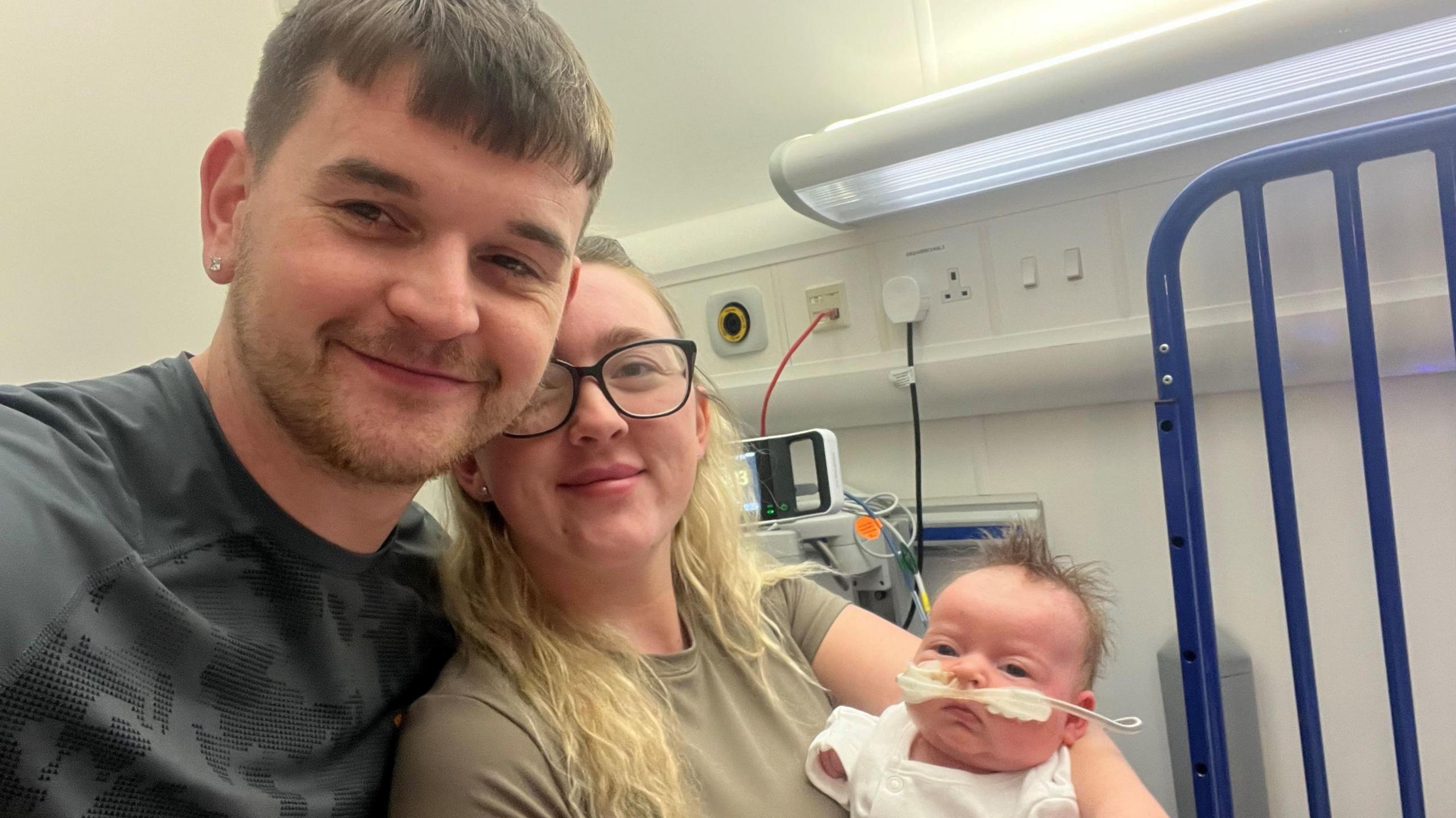 A woman and man smile at the camera, the woman holding a young baby. The baby is wearing a white babygrow and has a tube coming out of her nose, while medical equipment can be seen in the background.