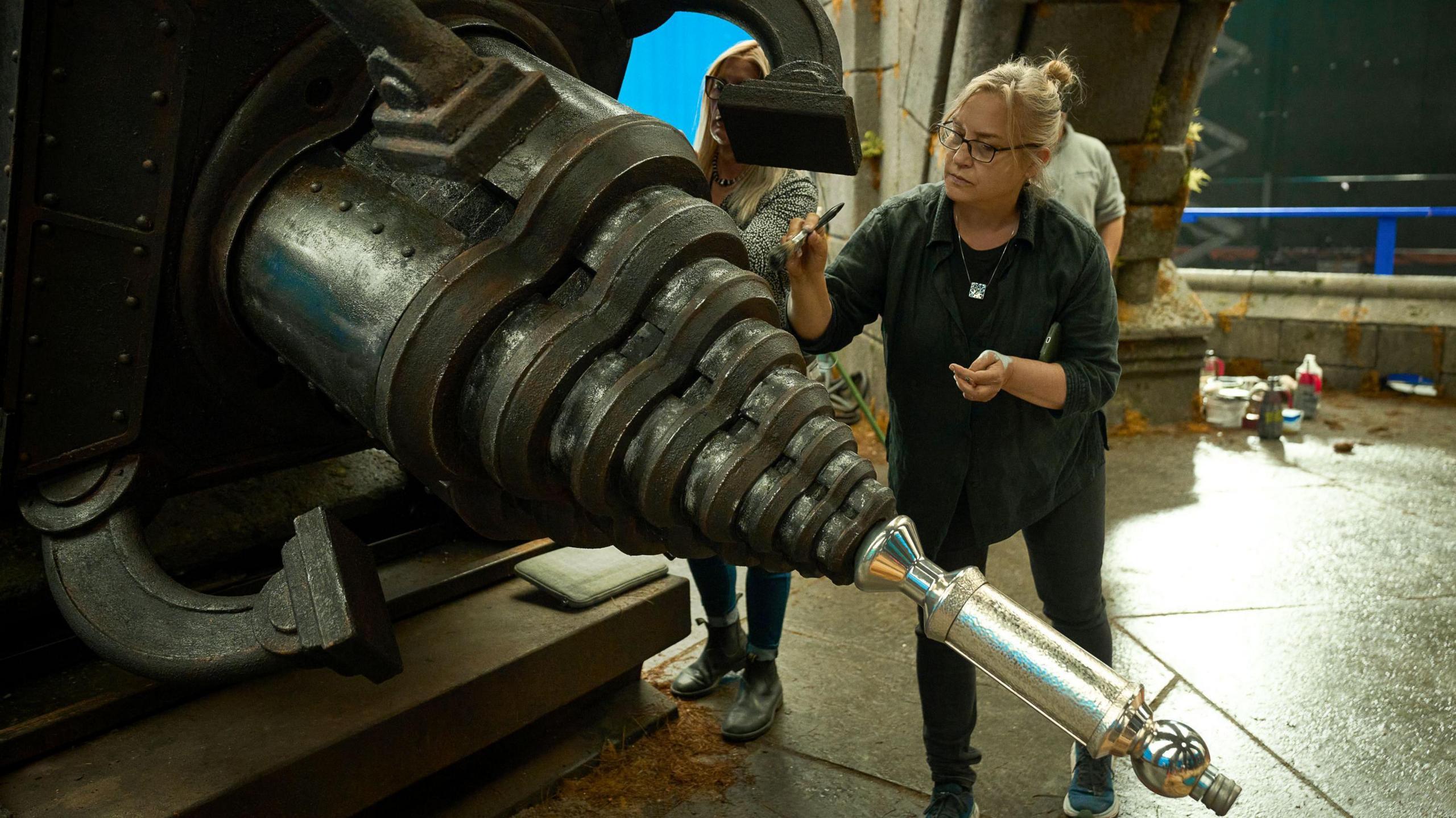 Tamara Deverell in green shirt and dark trousers using a brush to paint a pointy, steam-punk style prop which appears to be in a stone building.