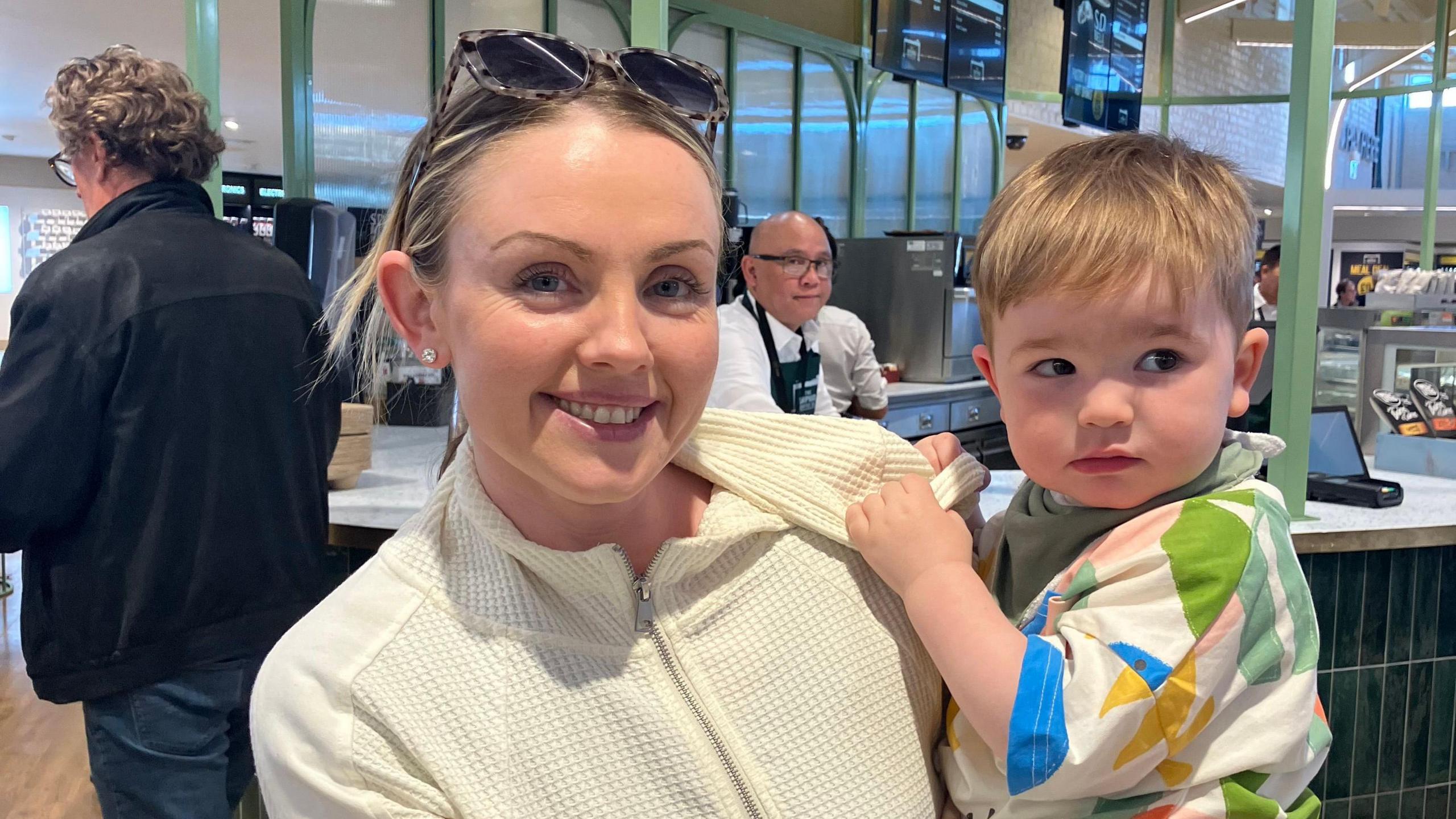 Judith is smiling at the camera wearing a white zip up and tortoise shell sunglasses on her hand. She has blond hair and blue eyes and is smiling at the camera. She has silver stud earrings in. She is holding her son who is blond with brown eyes. He is wearing a multi-coloured top and looking away from the camera. Behind them is a green and white counter with a waiter behind it.