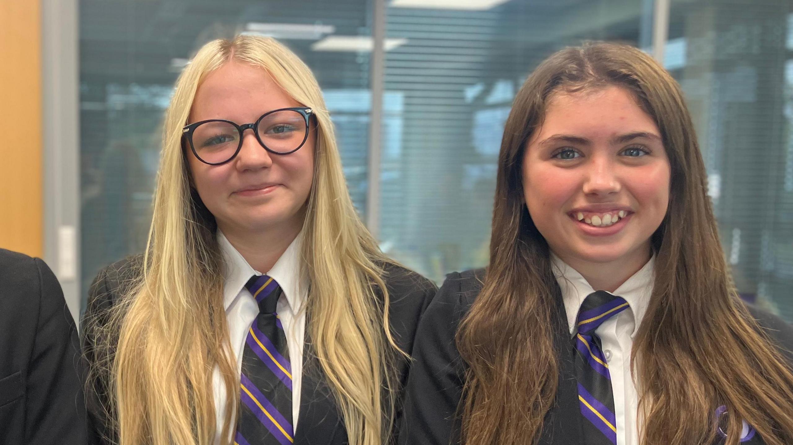Three young people smile at the camera. To the left a boy with dark hair wears a white shirt, black blazer and a tie. In the centre a girl with long blonde hair smiles wearing black spectacles, a black blazer and a shirt and tie. On the right, a girl with brunette hair smiles wearing a white shirt, a striped tie and a black blazer.