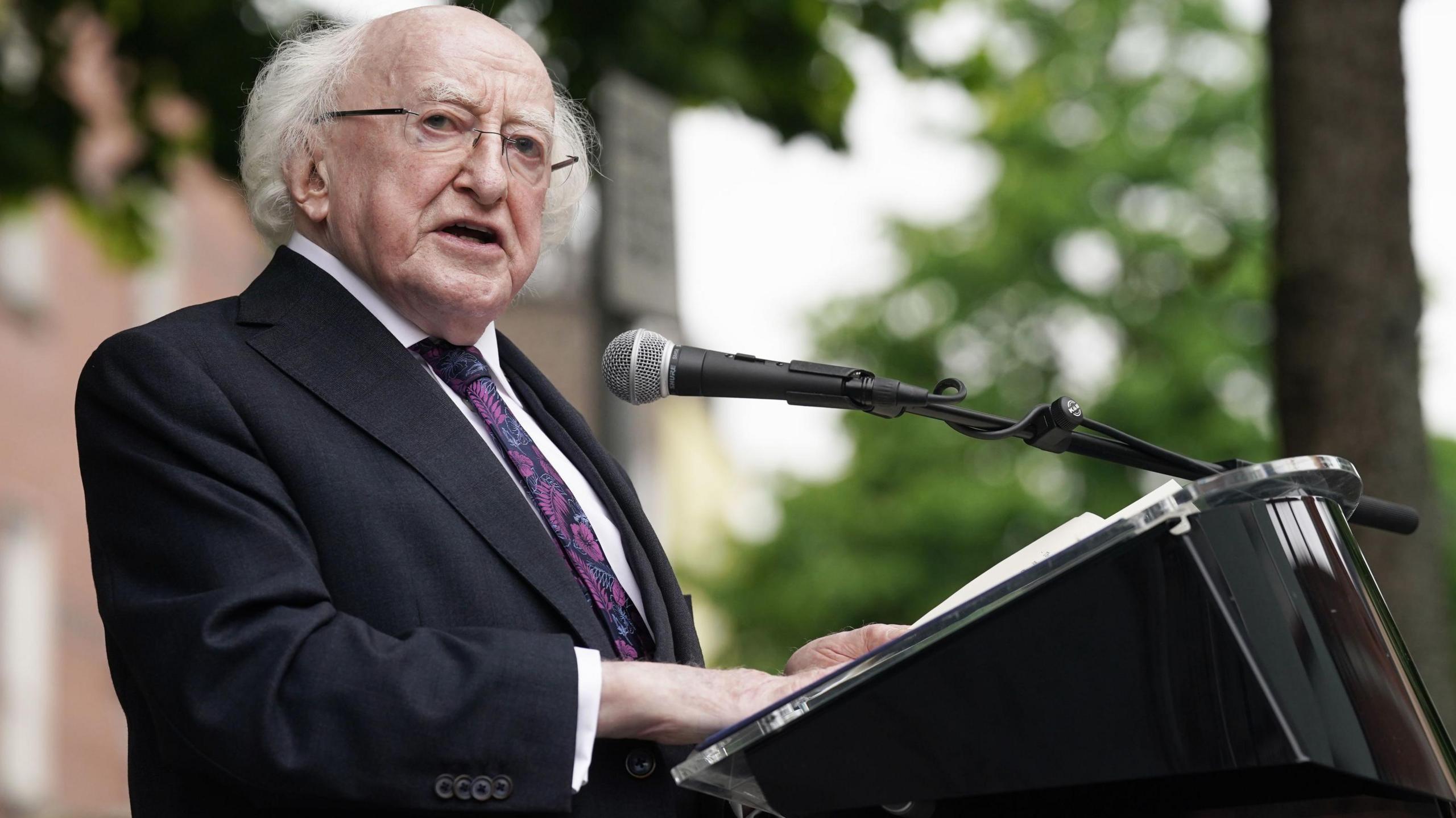 Michael D Higgins standing at a lectern he is wearing a black suit with a black tie that has purple flower patterns on it. He has glasses on.