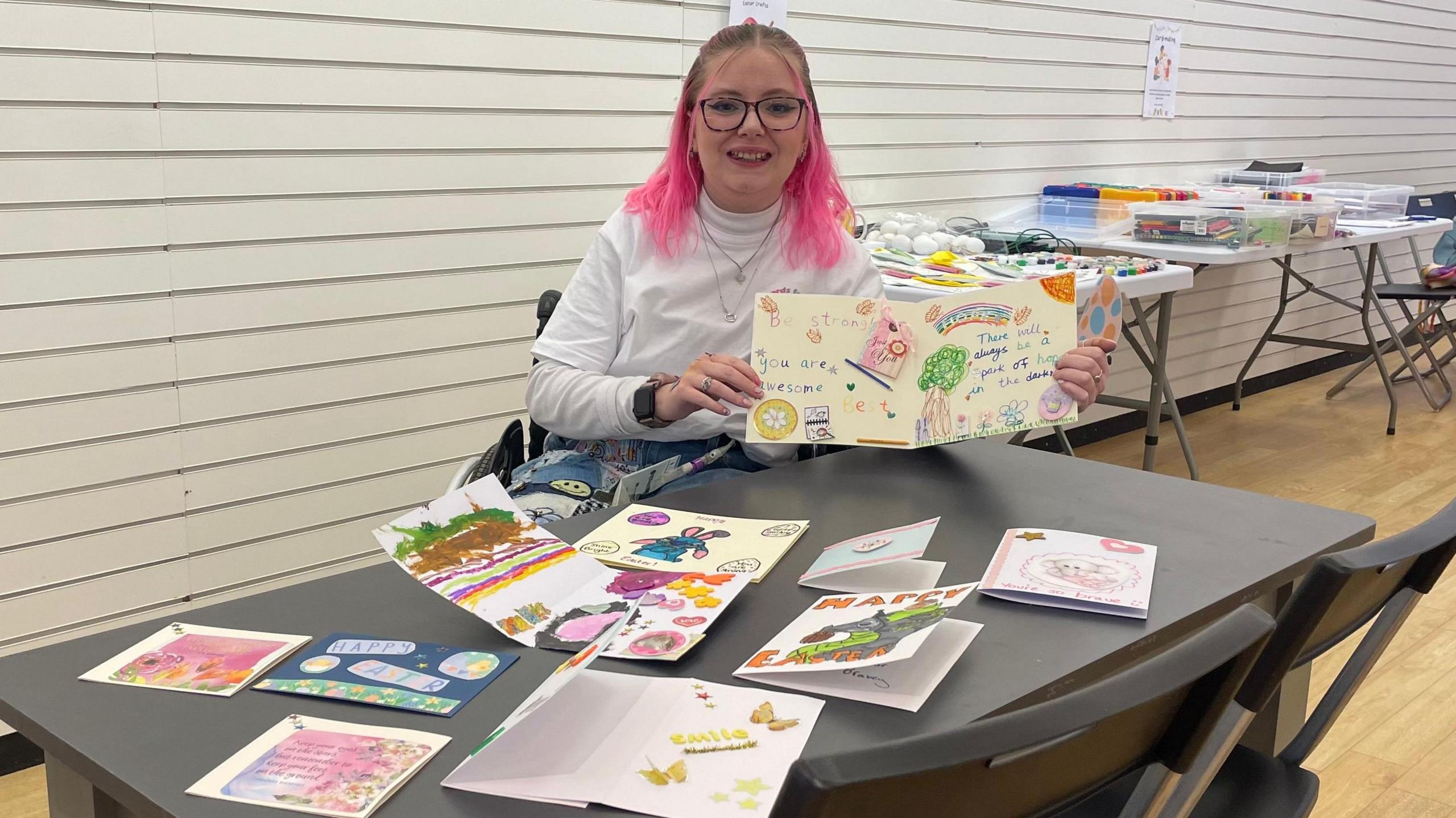 Katie Callaghan is wearing a white roll neck and has pink hair. She is sat at a grey table which is covered in handmade cards. She is holding one open towards the camera.