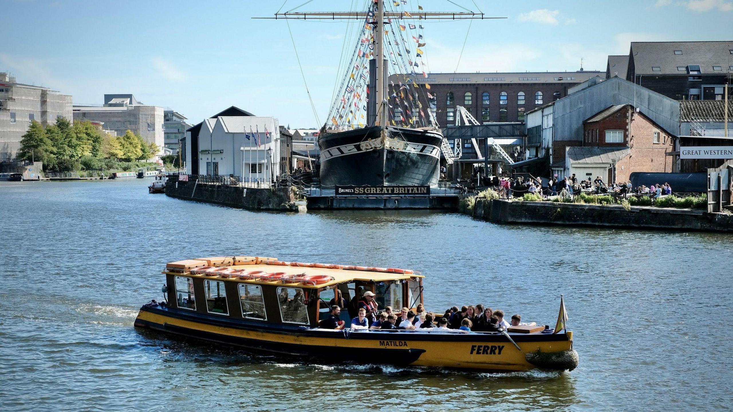 A Bristol Ferry Boat vessel travels across the city's harbour on a sunny day. It has many people on board and in the background the SS Great Britain can be seen in its dry dock