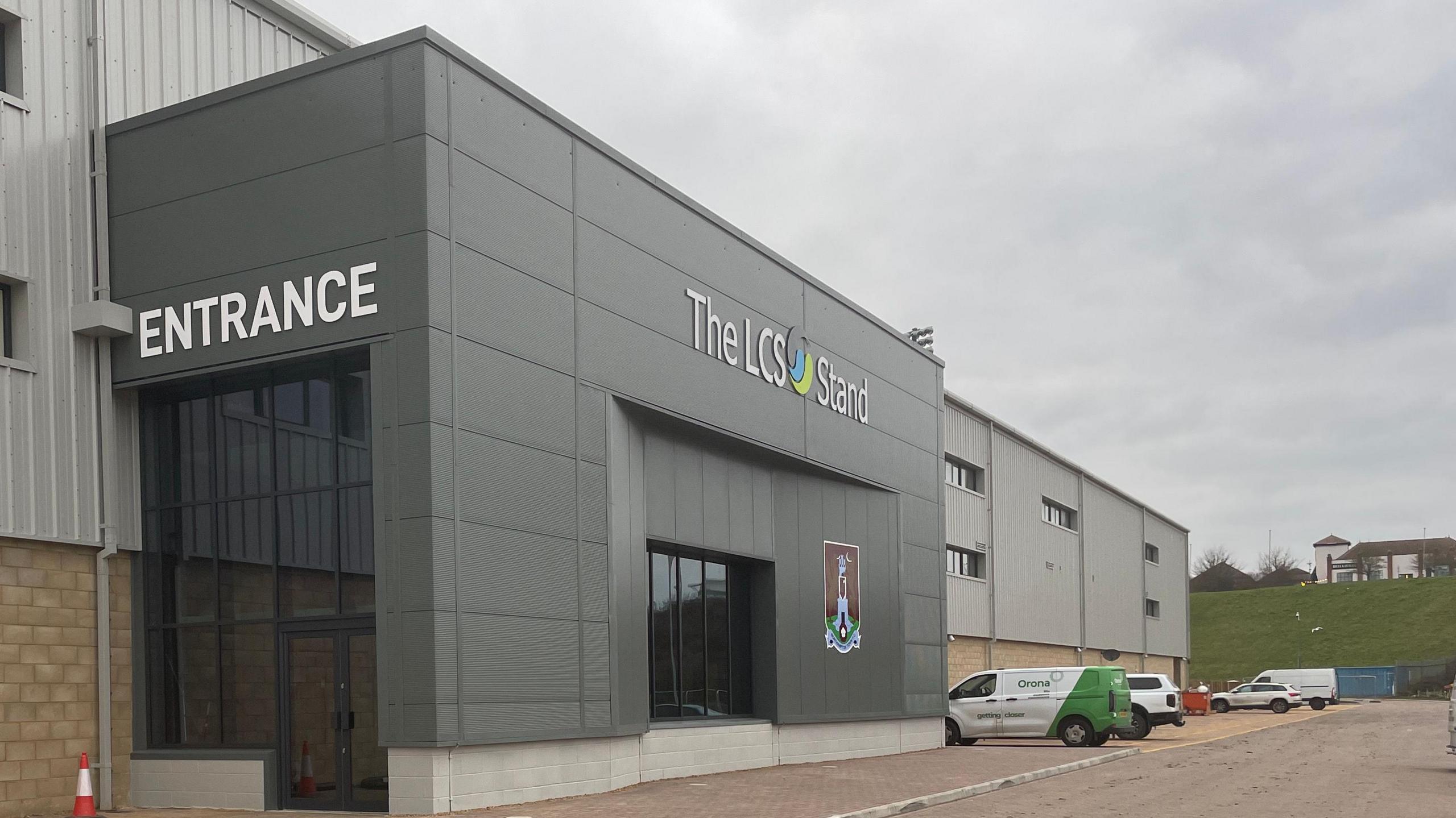 The entrance to the new stand featuring Northampton Town's crest on the left hand side of the entrance. Above the glass doors, a sign reading "The LCS Stand" can be seen. 