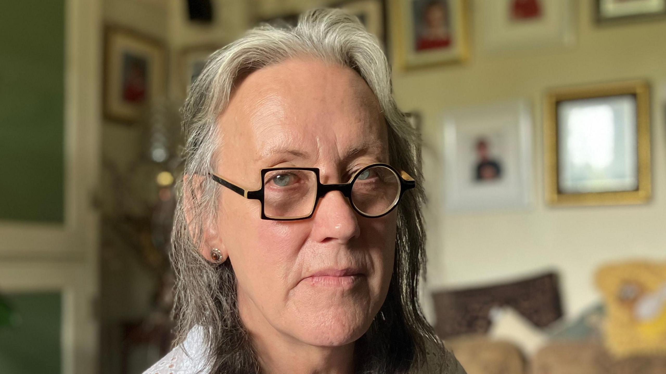 Ms Chaervey looks at the camera and she kneels in her living room. She wears black glasses. Behind her on the wall picture frames can be seen along with a sofa. 