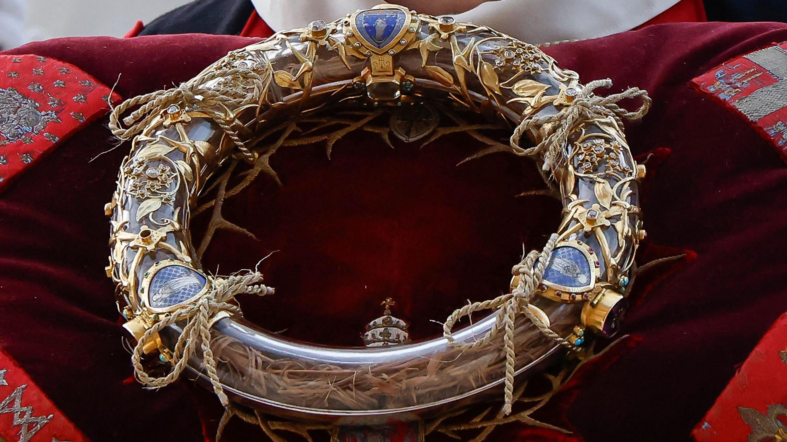 A close-up of the Crown of Thorns encased in a transparent tube with gold decorative details, placed on a red cushion. 