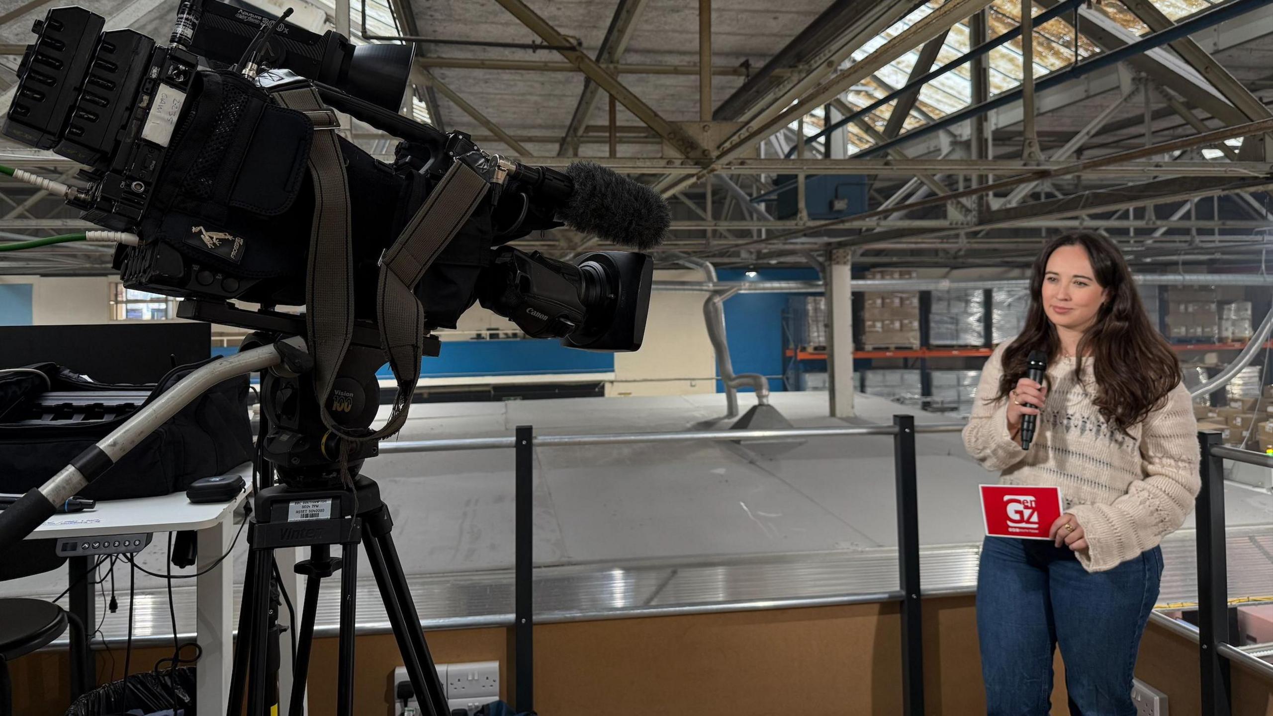 A woman with long black hair and wearing a cream-coloured jumper and skinny jeans. She's standing on the right hand side of the photo holding a microphone, with a camera pointed towards her. She appears to be standing inside a warehouse.