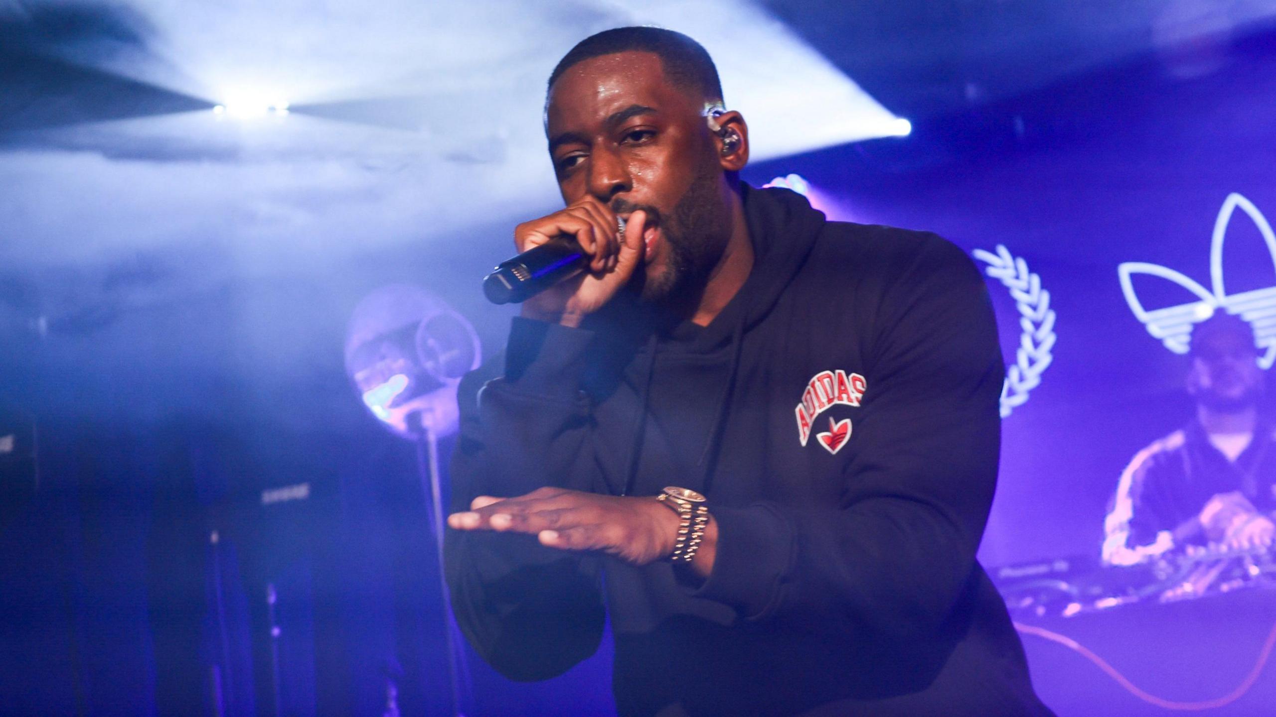 Bashy performing on stage in September 2024. He wears a black sports hoodie and stands in front of DJ decks, with the staging lit in purple. 