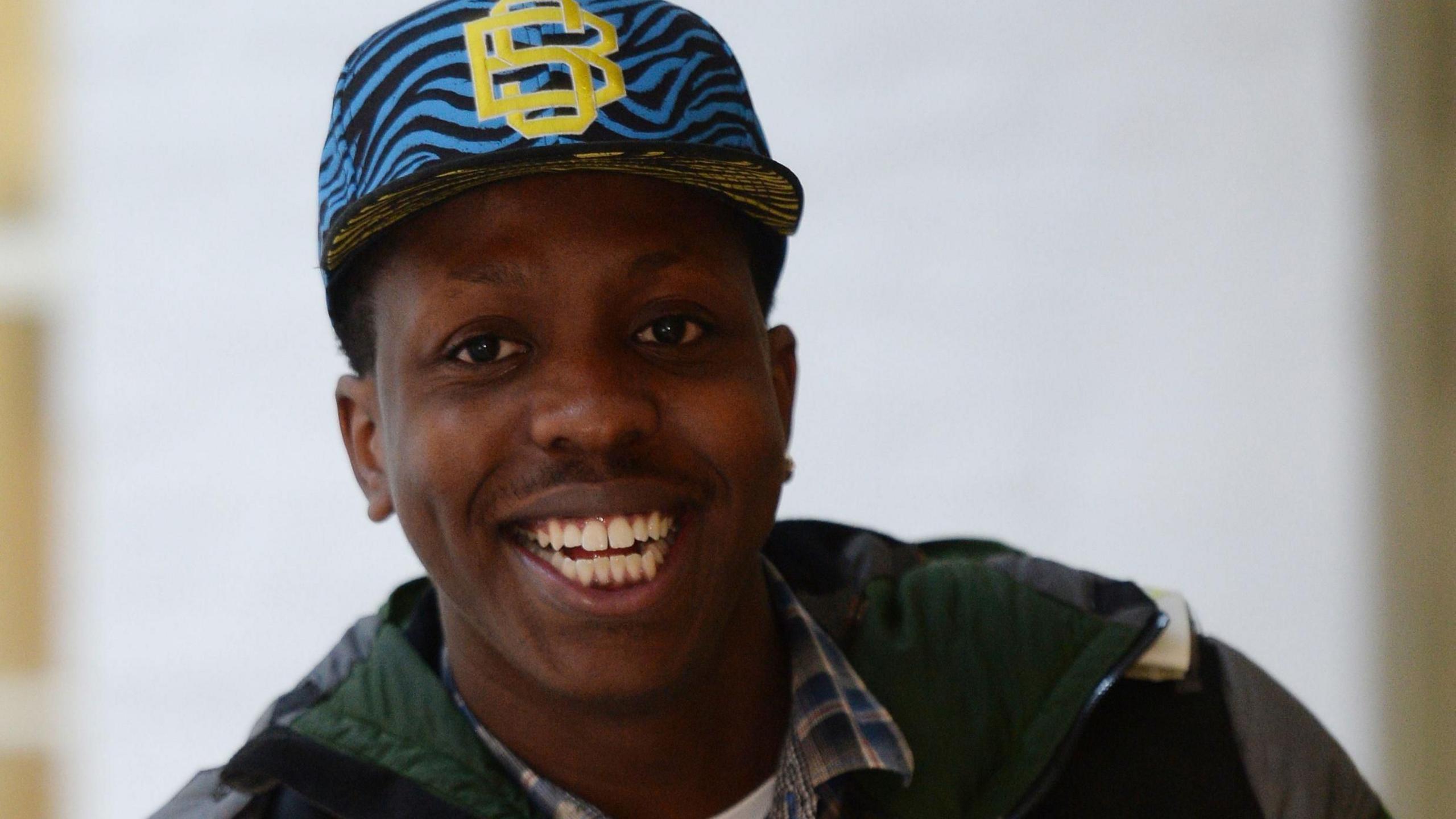 Jamal Edwards is smiling and wearing a blue baseball ca, a green coat and a blue check shirt.