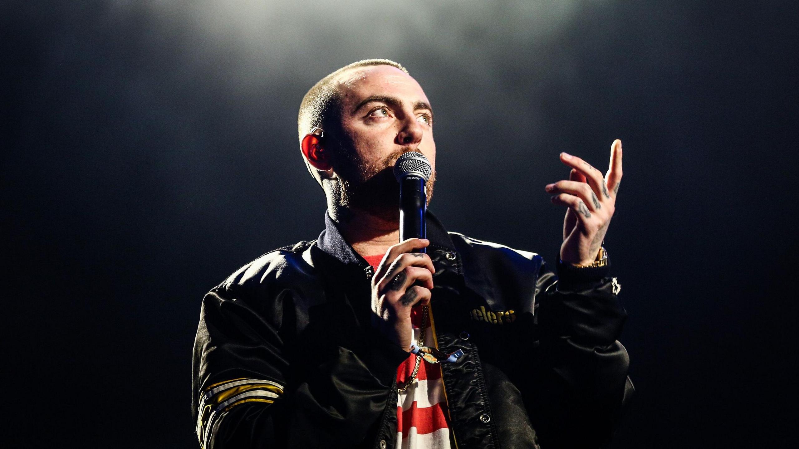 Mac Miller on a dark stage with a spotlight overhead. He wears a black bomber jacket over a red and white striped top and holds a mic to his face with his right hand while pointing skywards with his left. 