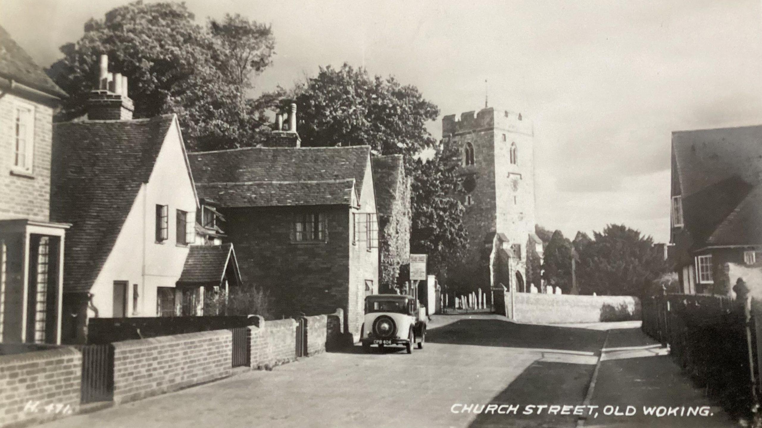 A black and white photograph of Church Street in Old Woking, showing a car parked outside a house and the church in the background.