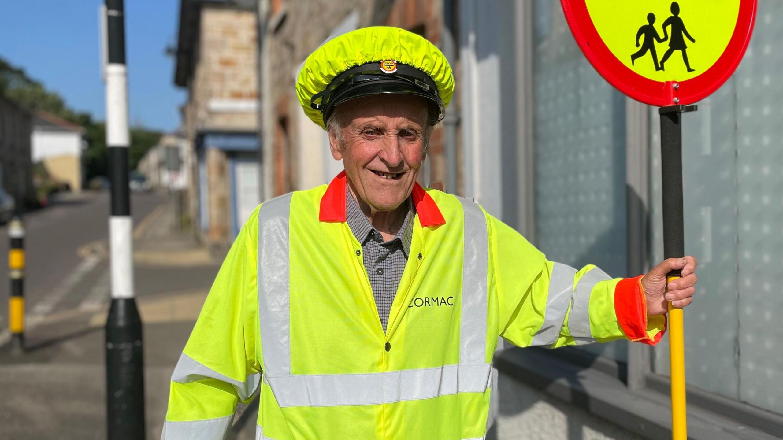 Richard Northey is in his lollipop man uniform which consists of a high-viz jacket and cap. He is smiling broadly at the camera as he stands in the street next to a zebra crossing and he is holding the lollipop which has a picture of two children walking hand in hand.