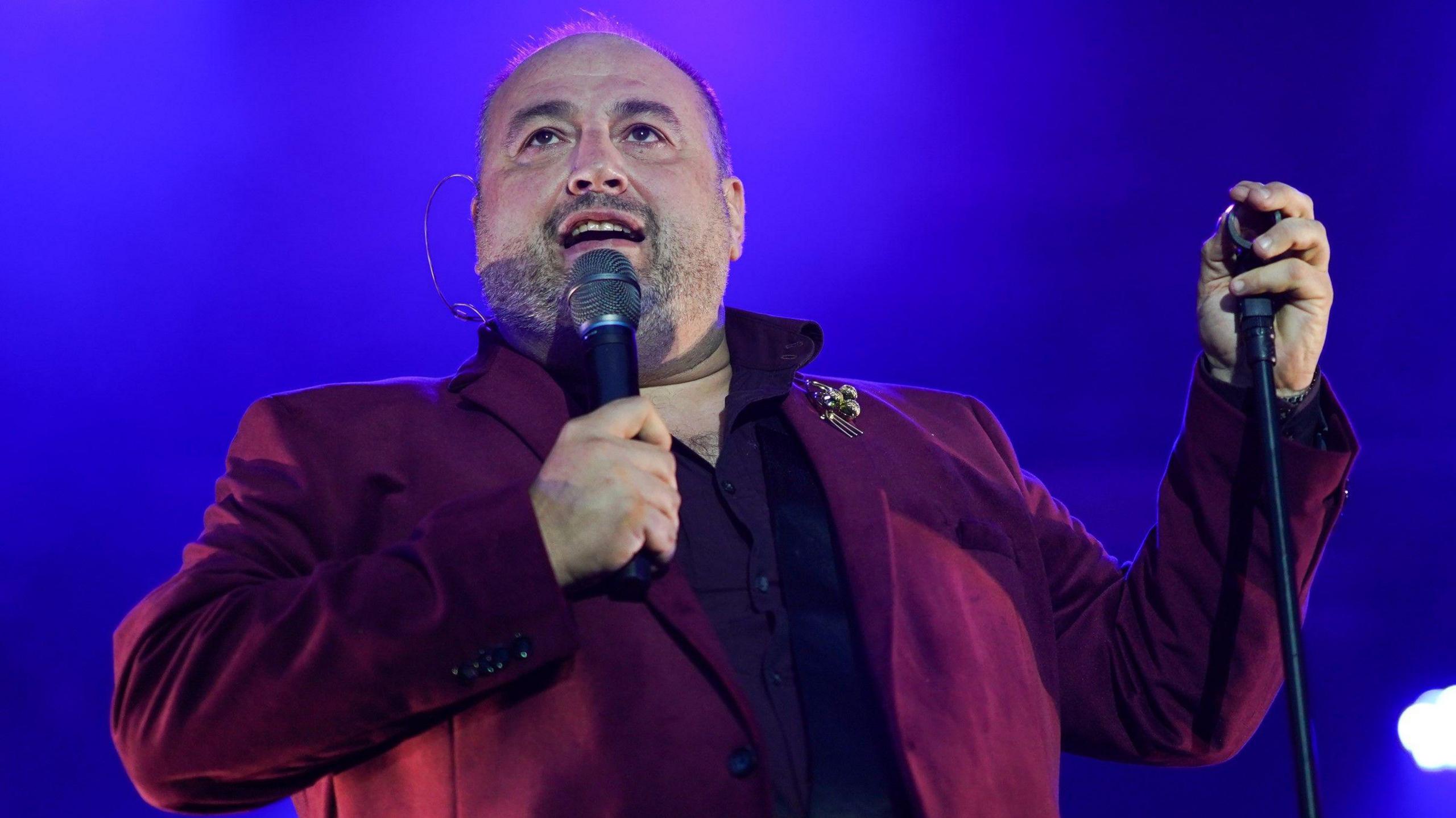 Wynne Evans, a middle-aged man, stands on stage in front of a blue background. He is wearing a burgundy suit and is holding a microphone