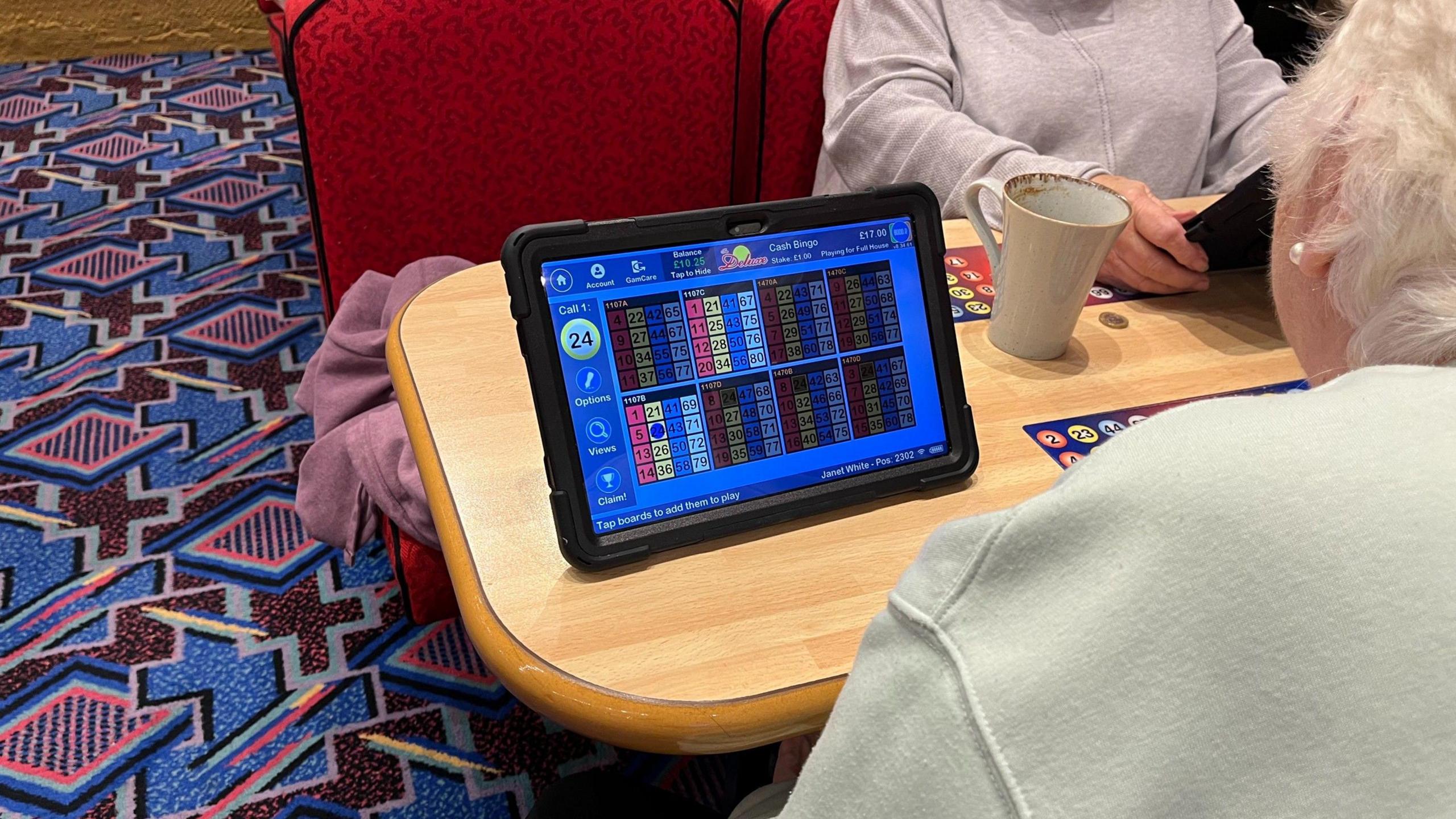 Two women site either side of a wooden table, playing bingo using electronic screens.