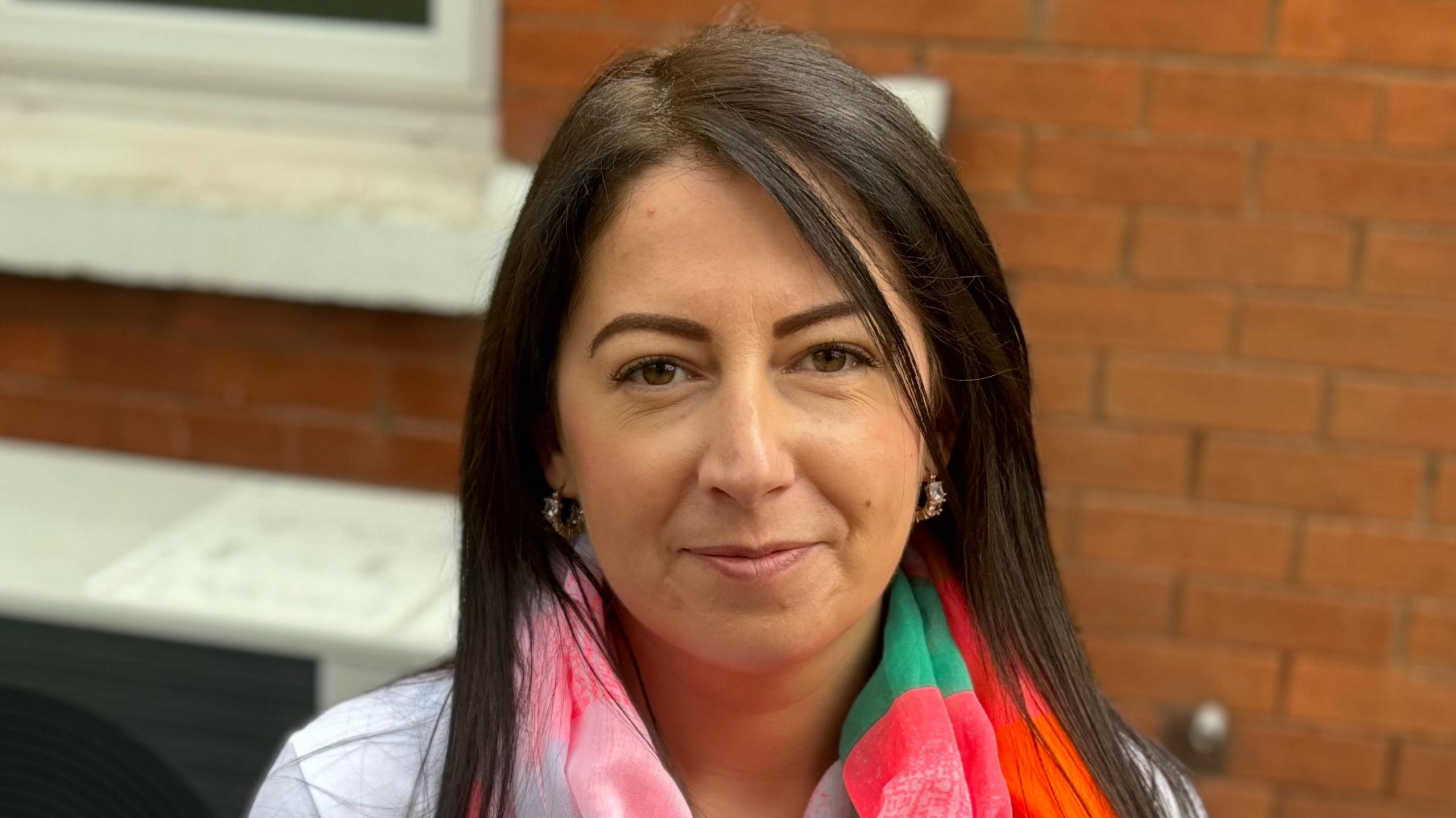 Stacey Fee, with long dark brown hair, stands in front of a red brick wall. She is wearing a white top and a coloured scarf