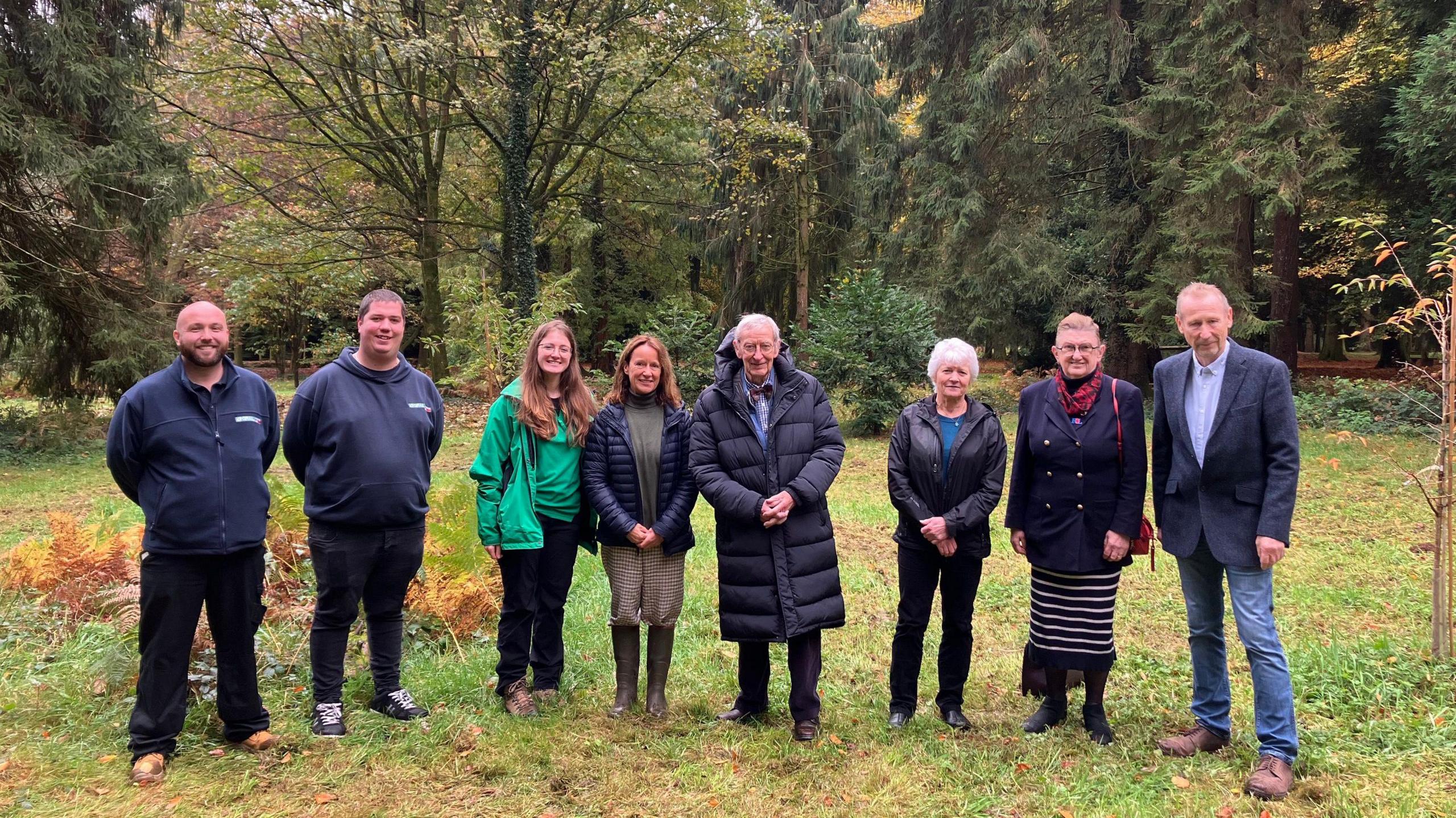 The Imjin Veterans Memorial Steering Group stand in a line at Cyril Hart Arboretum. The eight members are men and women of different ages. The trees and ferns that surround them in the clearing they are stood in are browning as it is autumn.