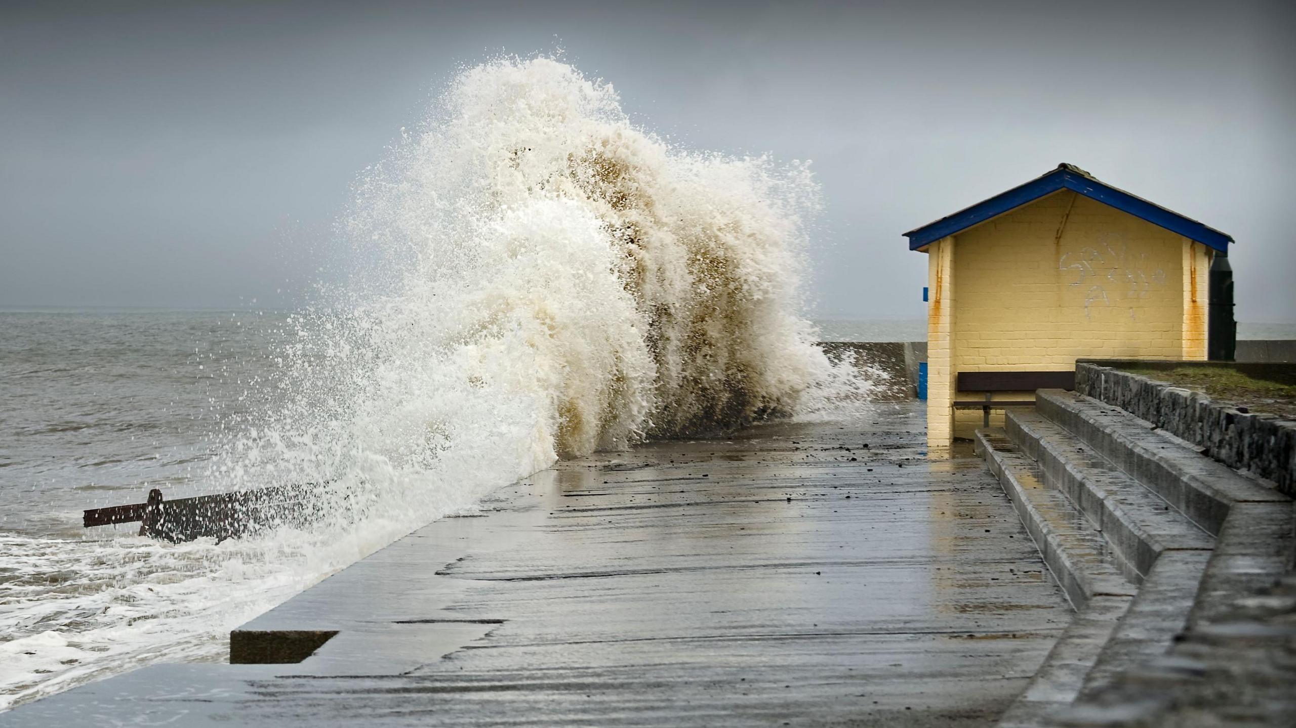 A wave crashes on a promenade. There is a hut with a bench inside near the wave. The clouds are grey and stormy.