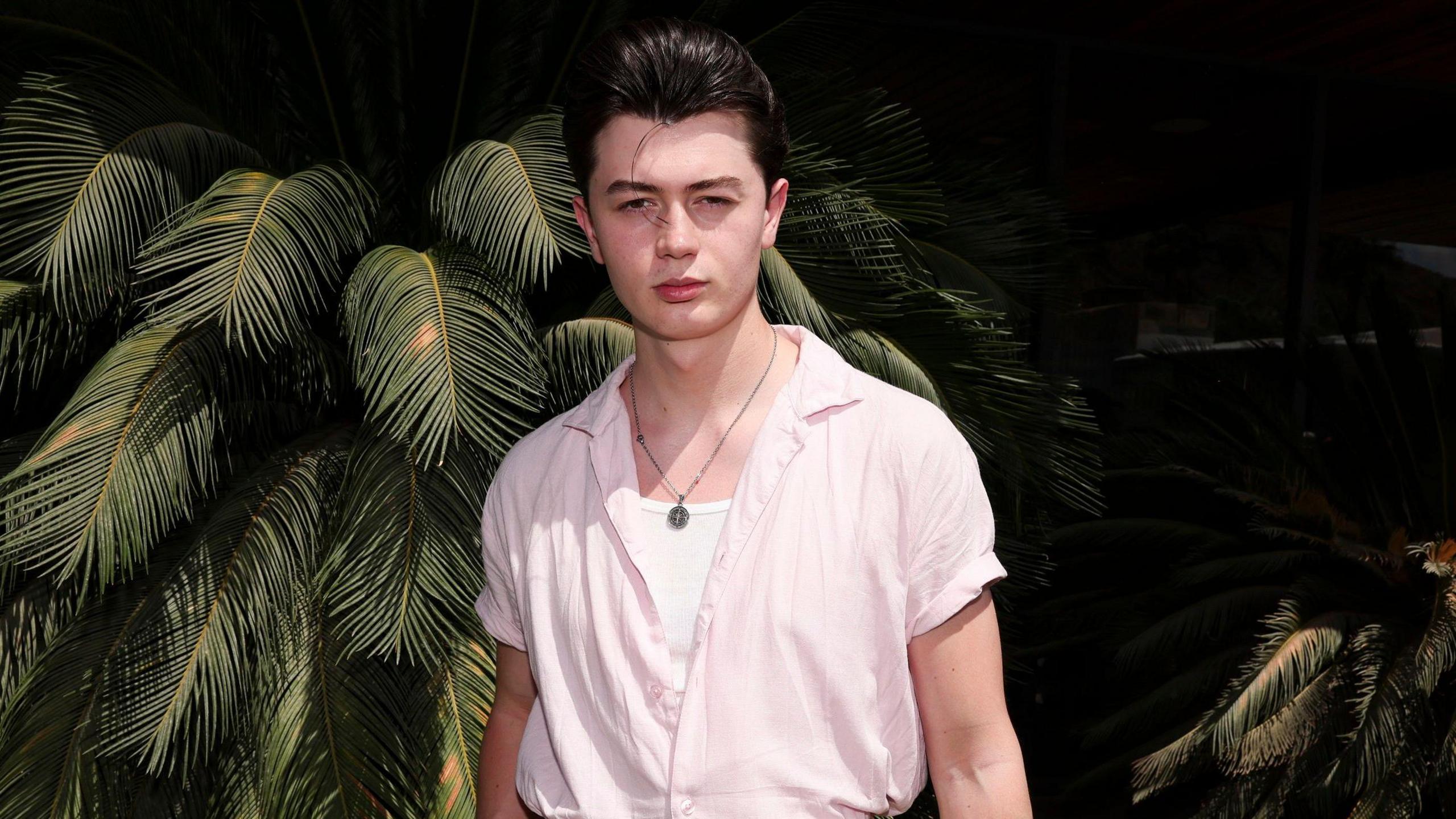 Reay poses at a California party in front of a palm tree plant. He has a dark quiff and wears a short sleeved pink shift over a white top and chain