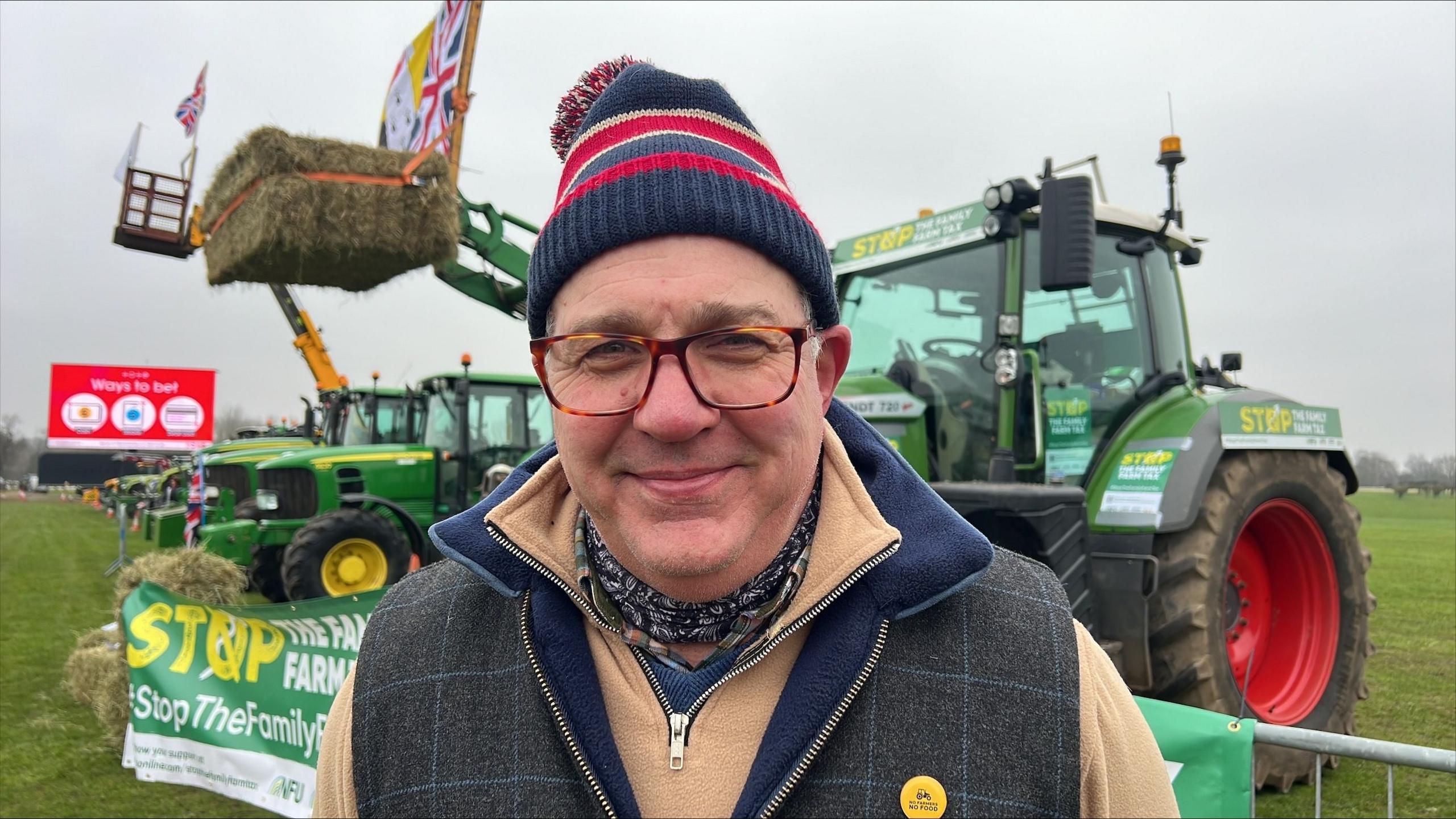 Tim Bradshaw wearing a read and blue bobble hat, patterned neckerchief, a beige zipped jumper and a country-style zipped gillet. He is standing in front of a tractor which has lifted a hay bale.