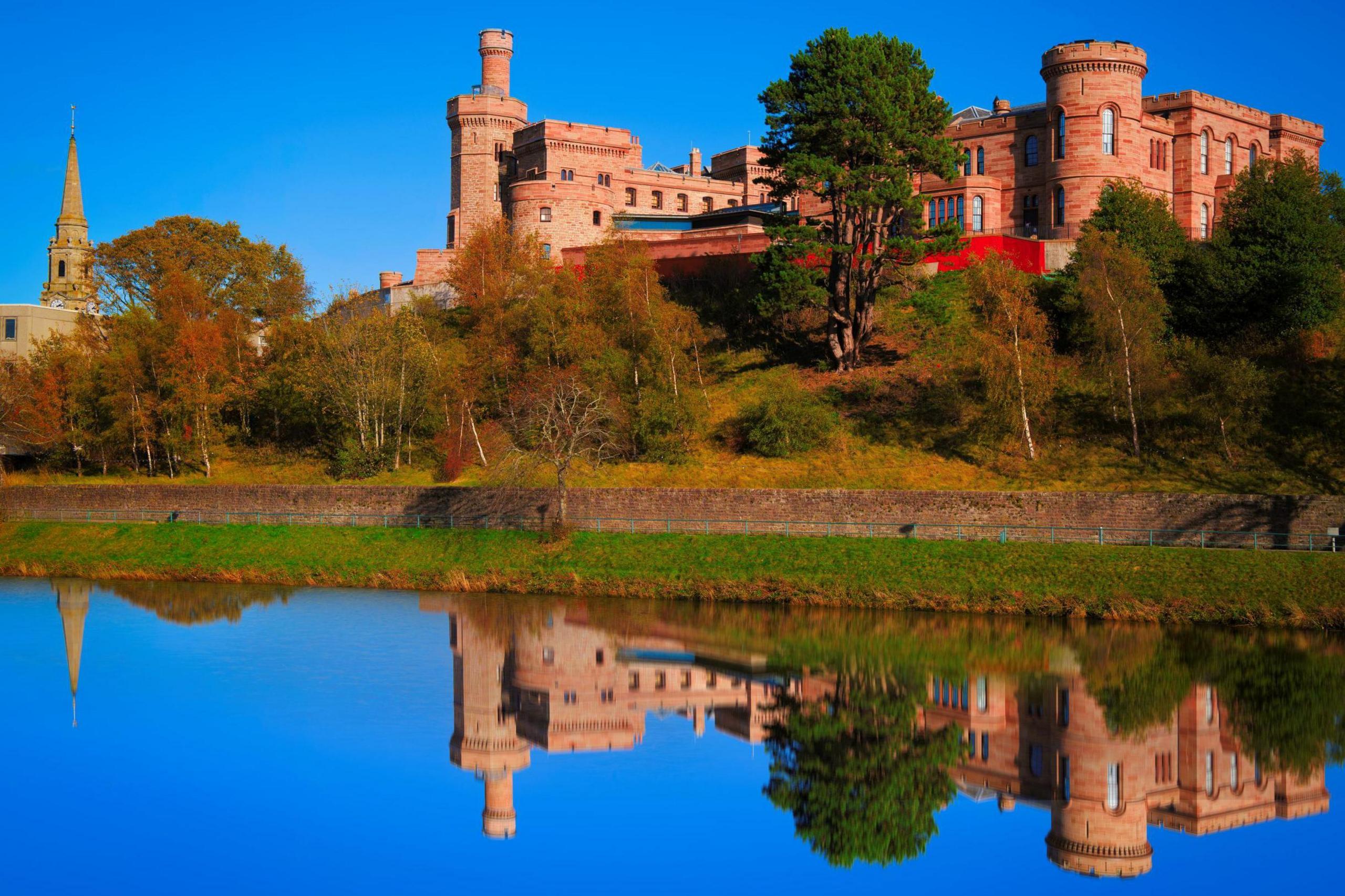 Inverness Castle is a red sandstone building with mock turrets and battlements. The castle is reflected in the mirror-like water of the river below.