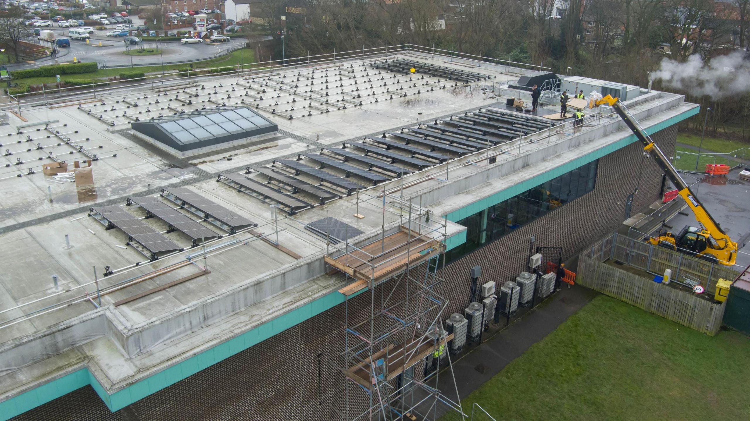 The picture shows the roof and construction under way. A row of solar panels is already installed. A yellow cherry picker is pictured on the right, with several people stood in front of it on the roof.