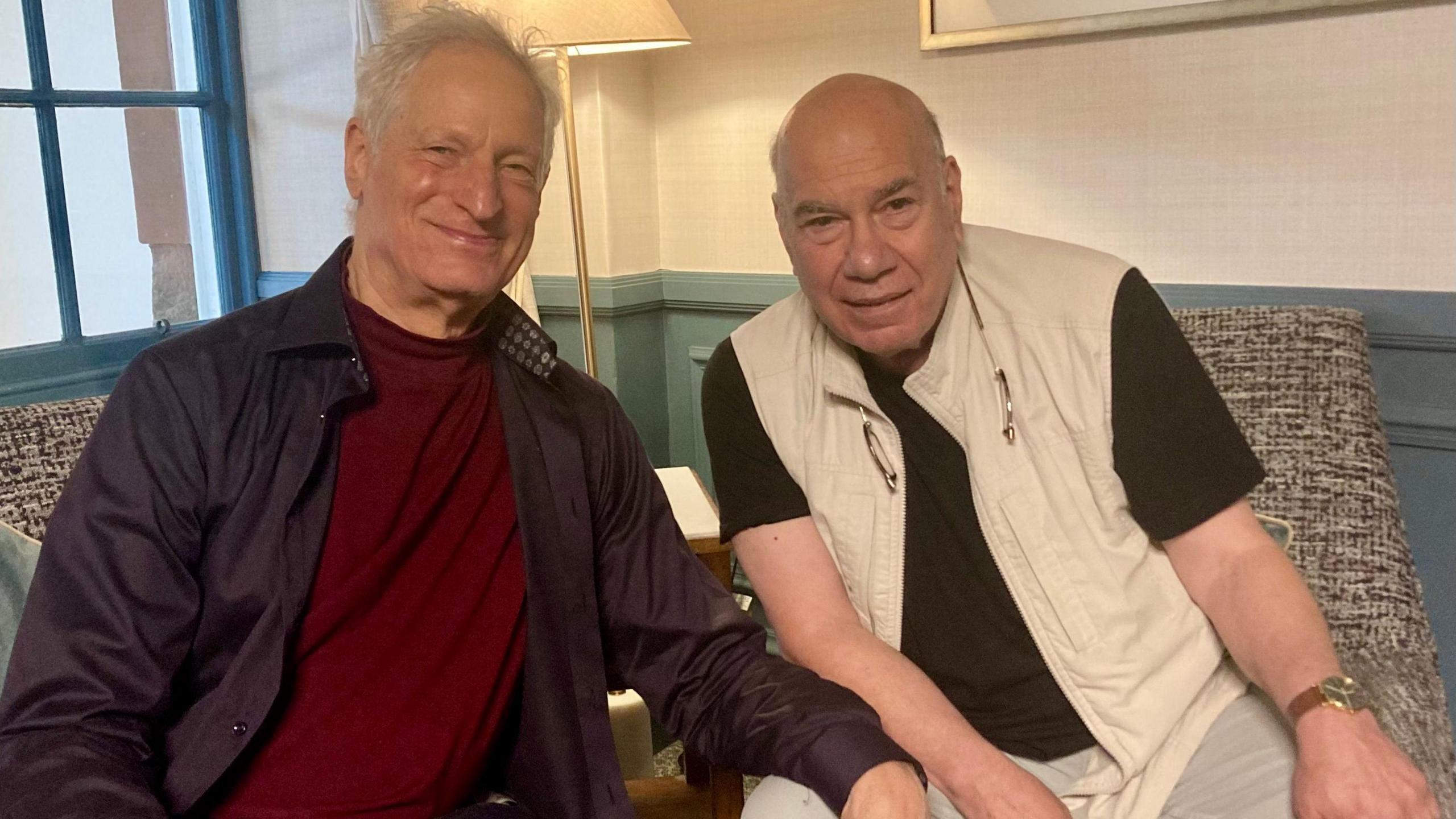 Marc Turtletaub has white hair and is wearing a brown shirt and maroon T-shirt. He is sitting on a chair in a well-appointed room. He is sitting next to Jay Parini, a bald man in a black T-shirt and cream gilet. They are smiling.