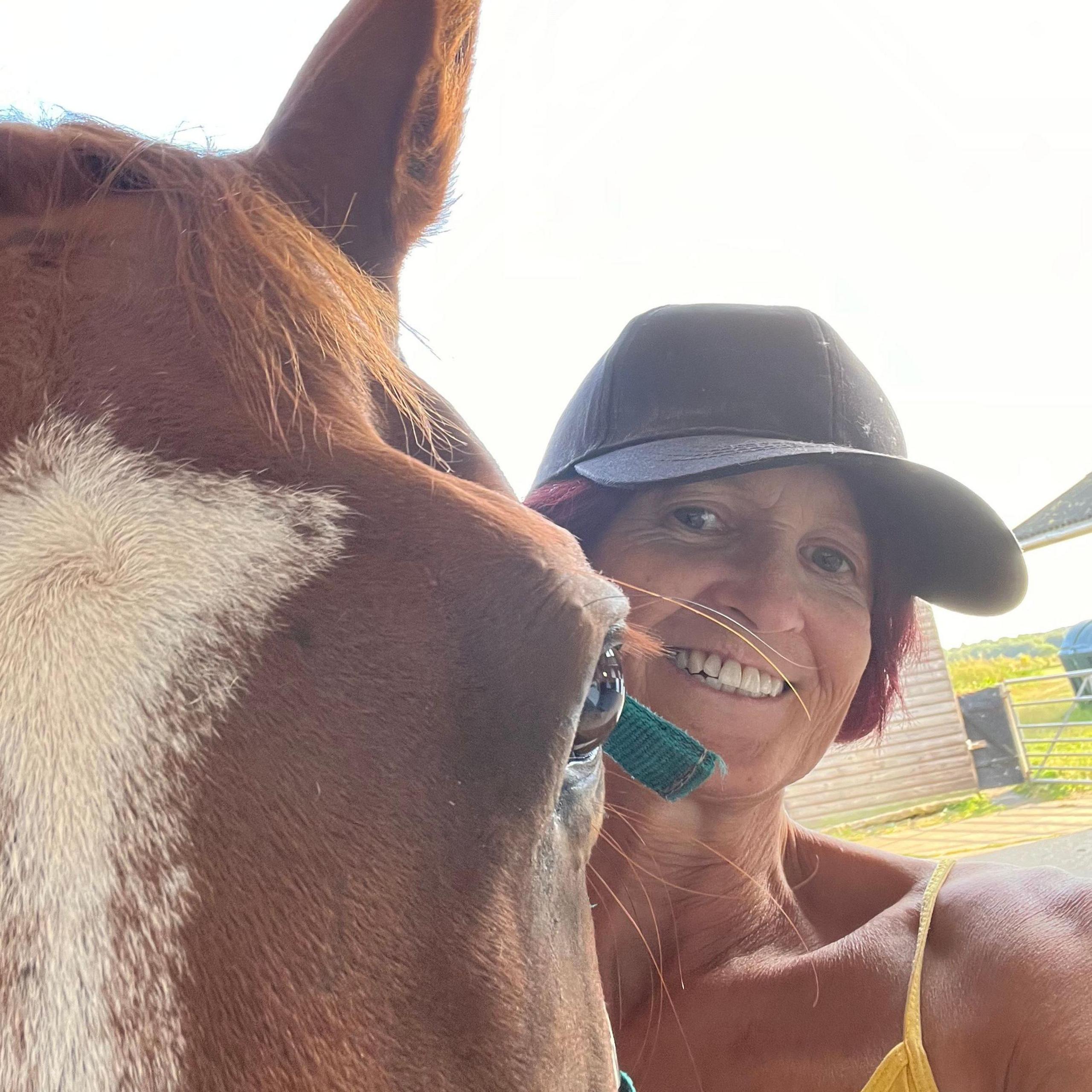 Sue - a woman smiling while wearing a baseball cap - stands next to a horse. The horse is closer to the camera, with only it's eye and forehead visible beside her.