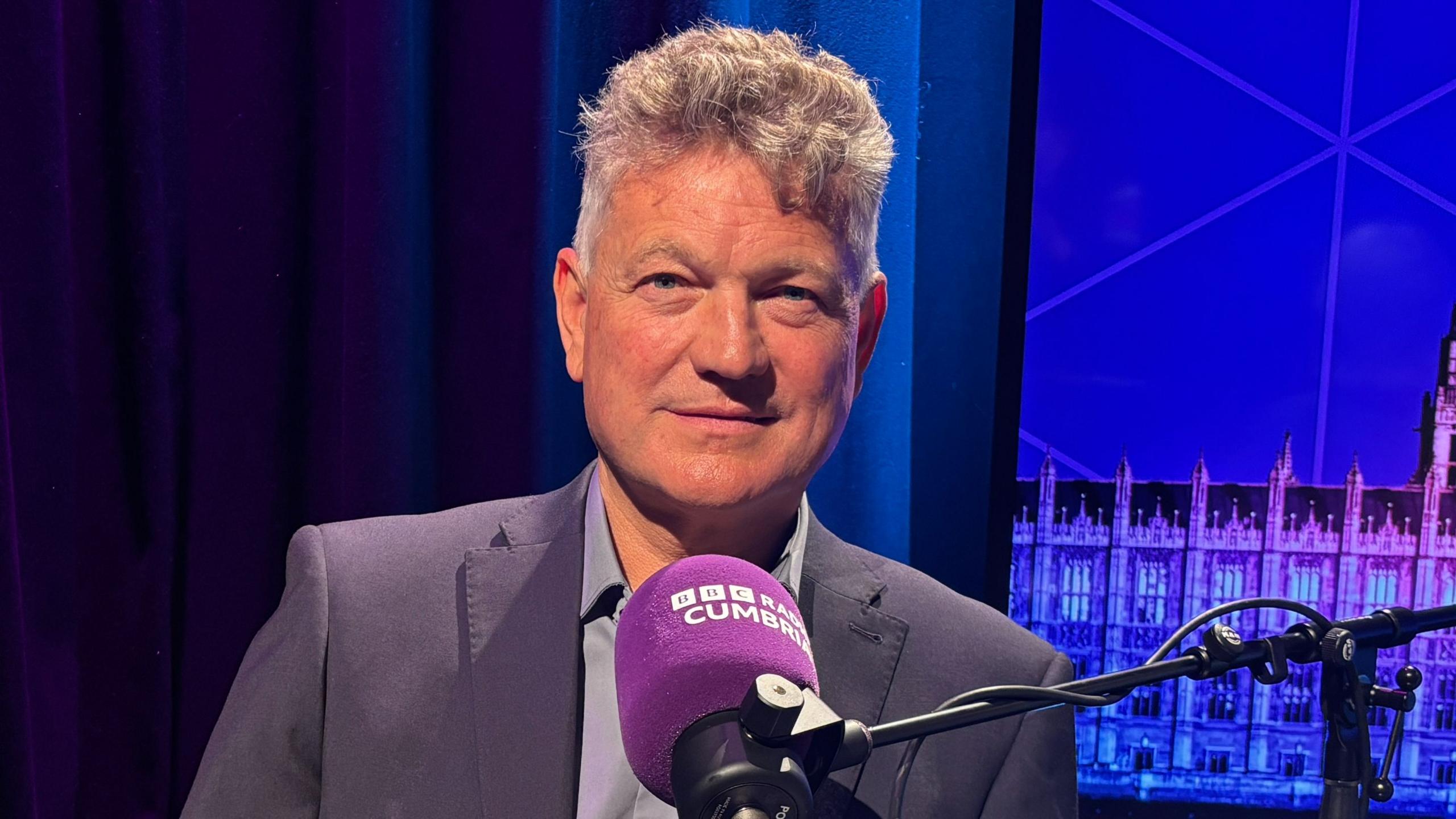 Mike Starkie sitting in front of a Radio Cumbria branded microphone. He's wearing a grey/purple suit and has short grey hair. 