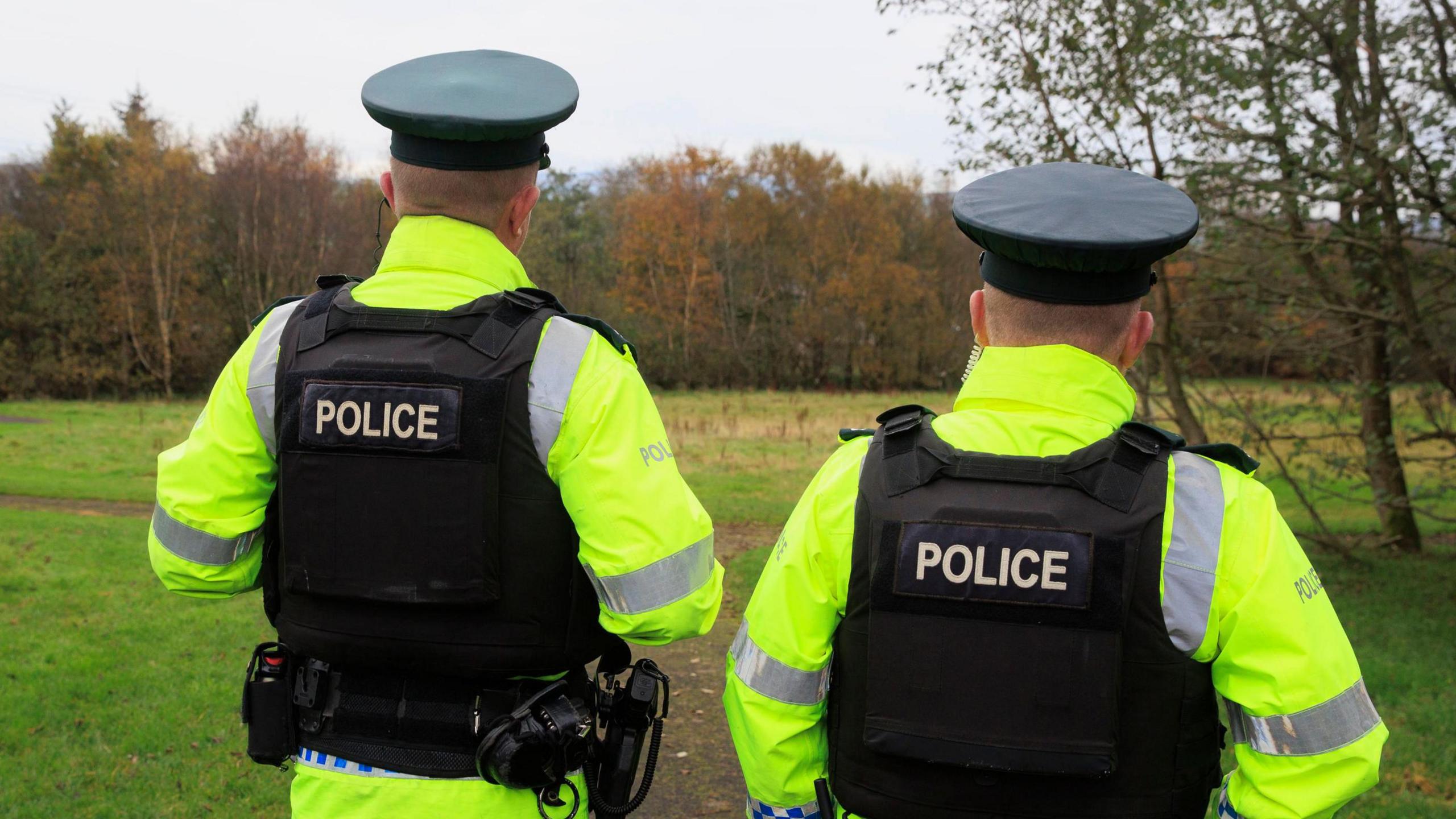 Landscape image of two police officers with their backs turned to the camera, they are wearing black vests with police written on the back and yellow hi-vis jackets. They are standing in a field.