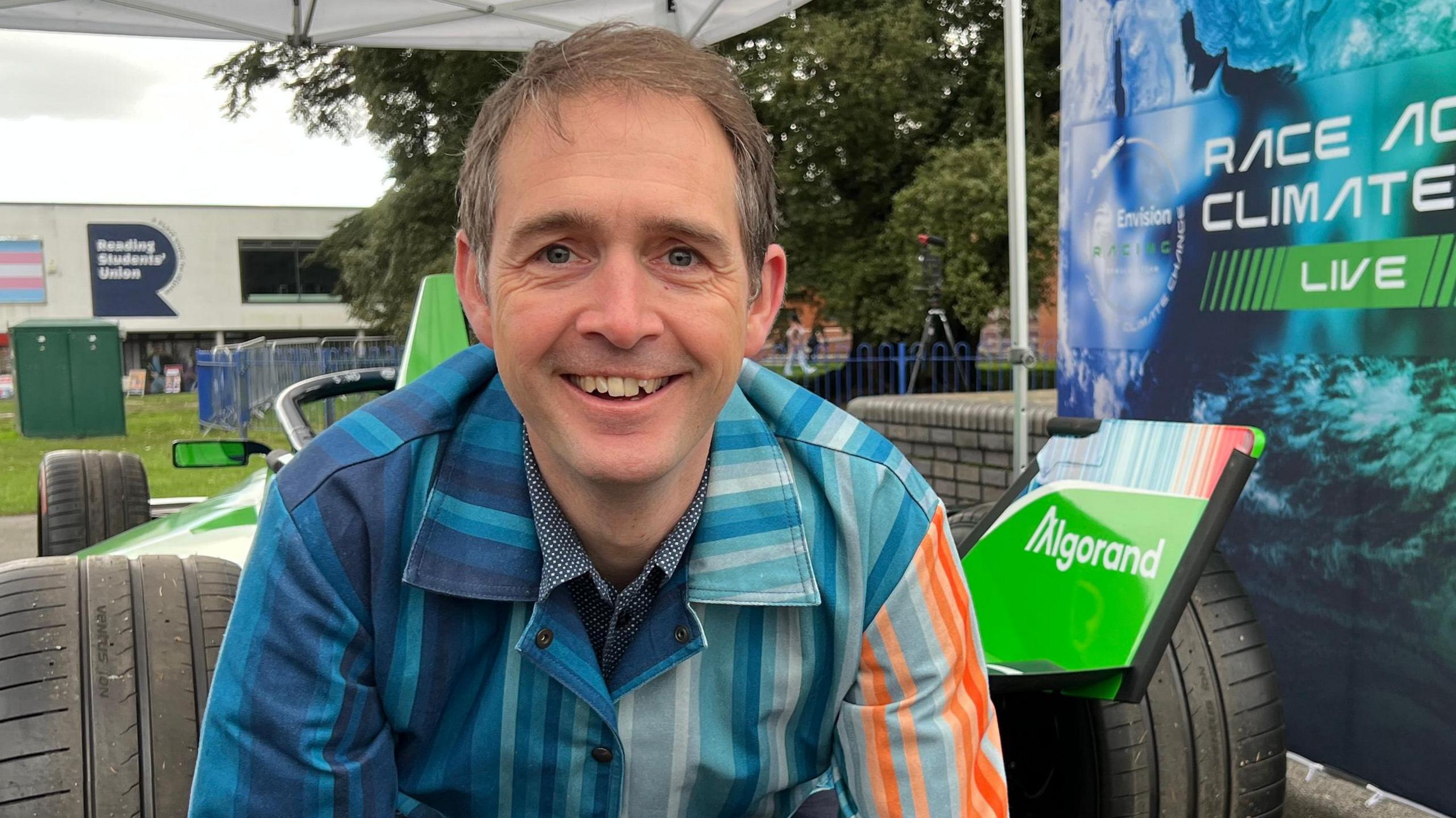 Ed has short brown hair, he is kneeling by the back of the car. He is wearing a striped suit in the colours of the climate stripes. Behind him is the rear wing of the car featuring his design.