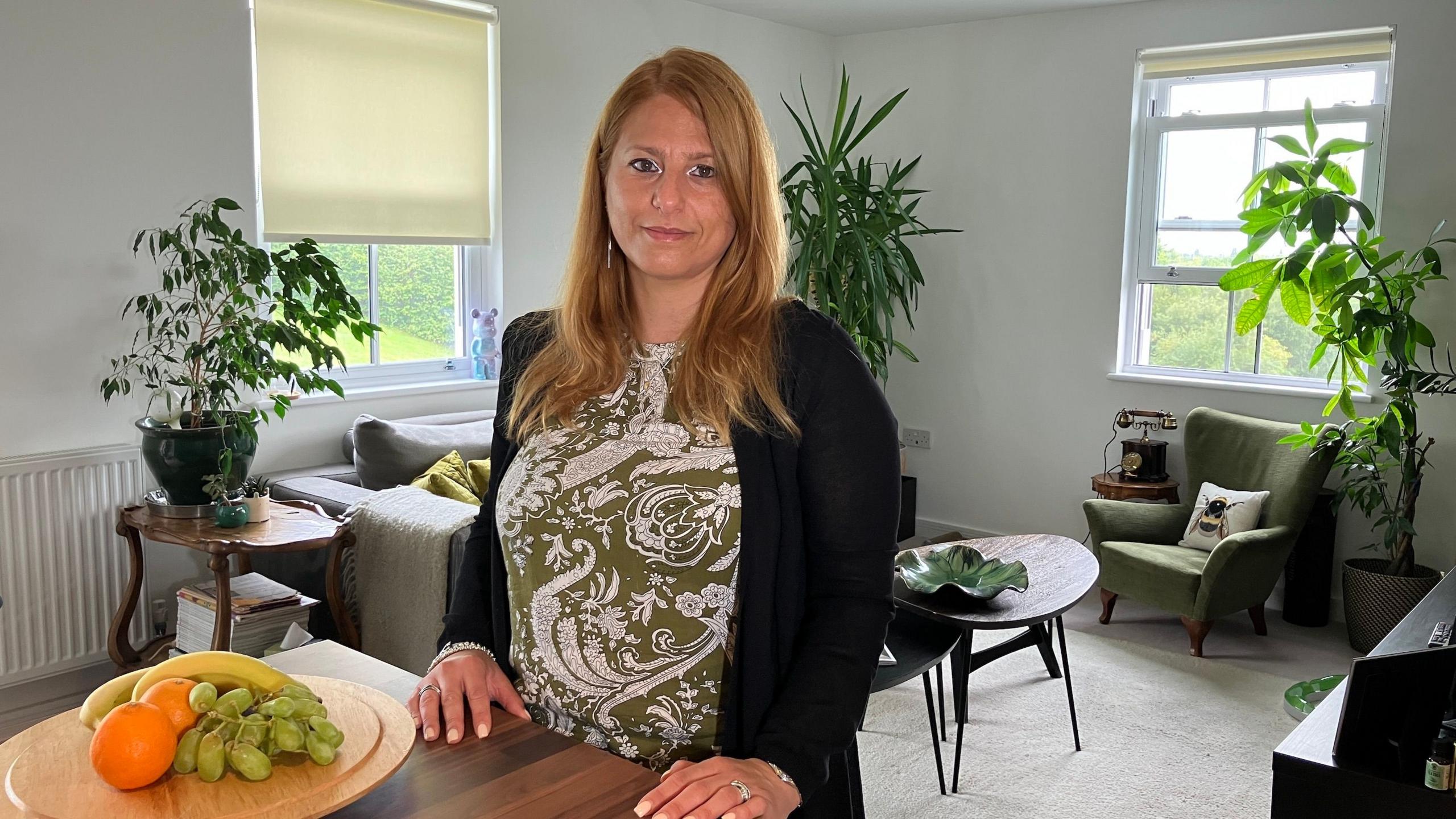 A woman with strawberry-blonde hair, wearing an olive green and white patterned top and black cardigan looks into the camera from her living room with plants in the background, a coffee table with large leaf-shaped dish and a plate of fruit in the foreground