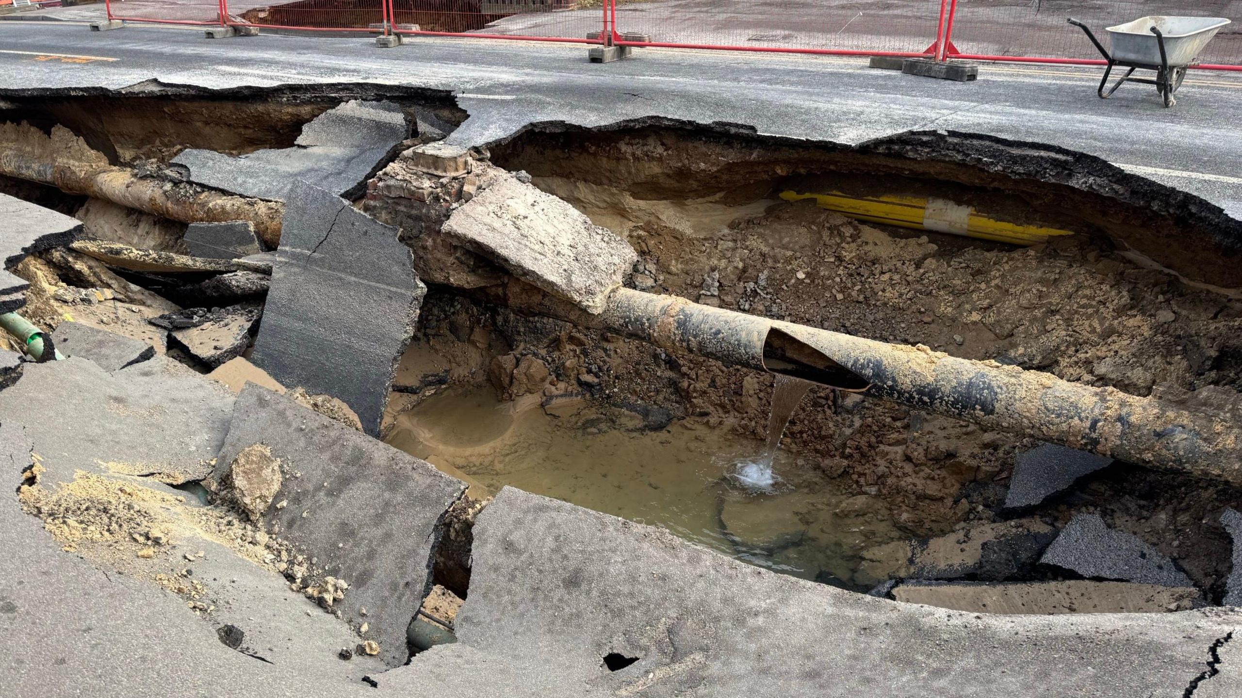 A giant hole in the road with a pipe running through it. There is murky water spilling from the pipe into the hole.