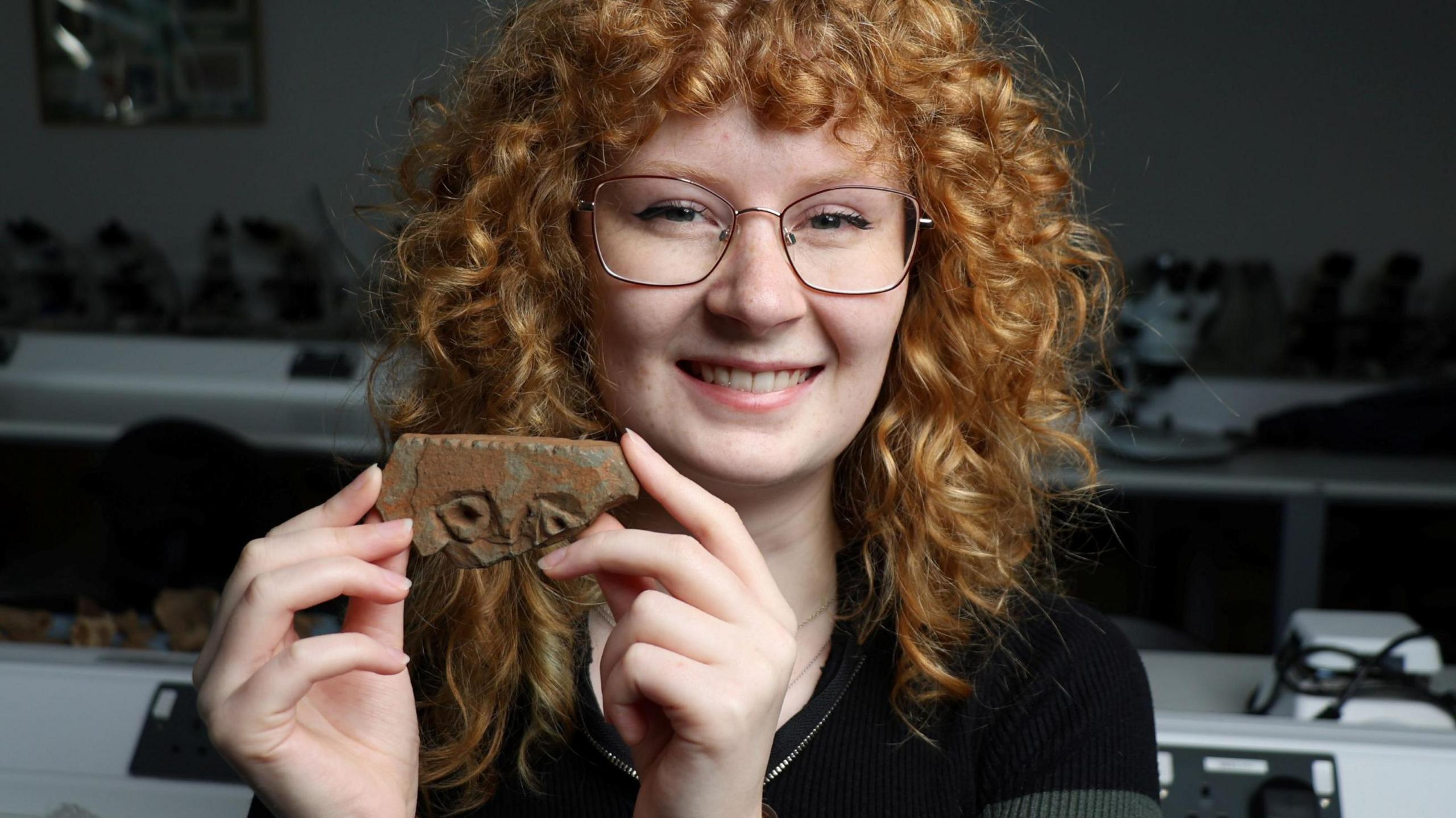 Jodie Allan is inside a university lab. She has long, curly red hair and is wearing glasses. She is smiling as she holds up the fragment of stone carving.