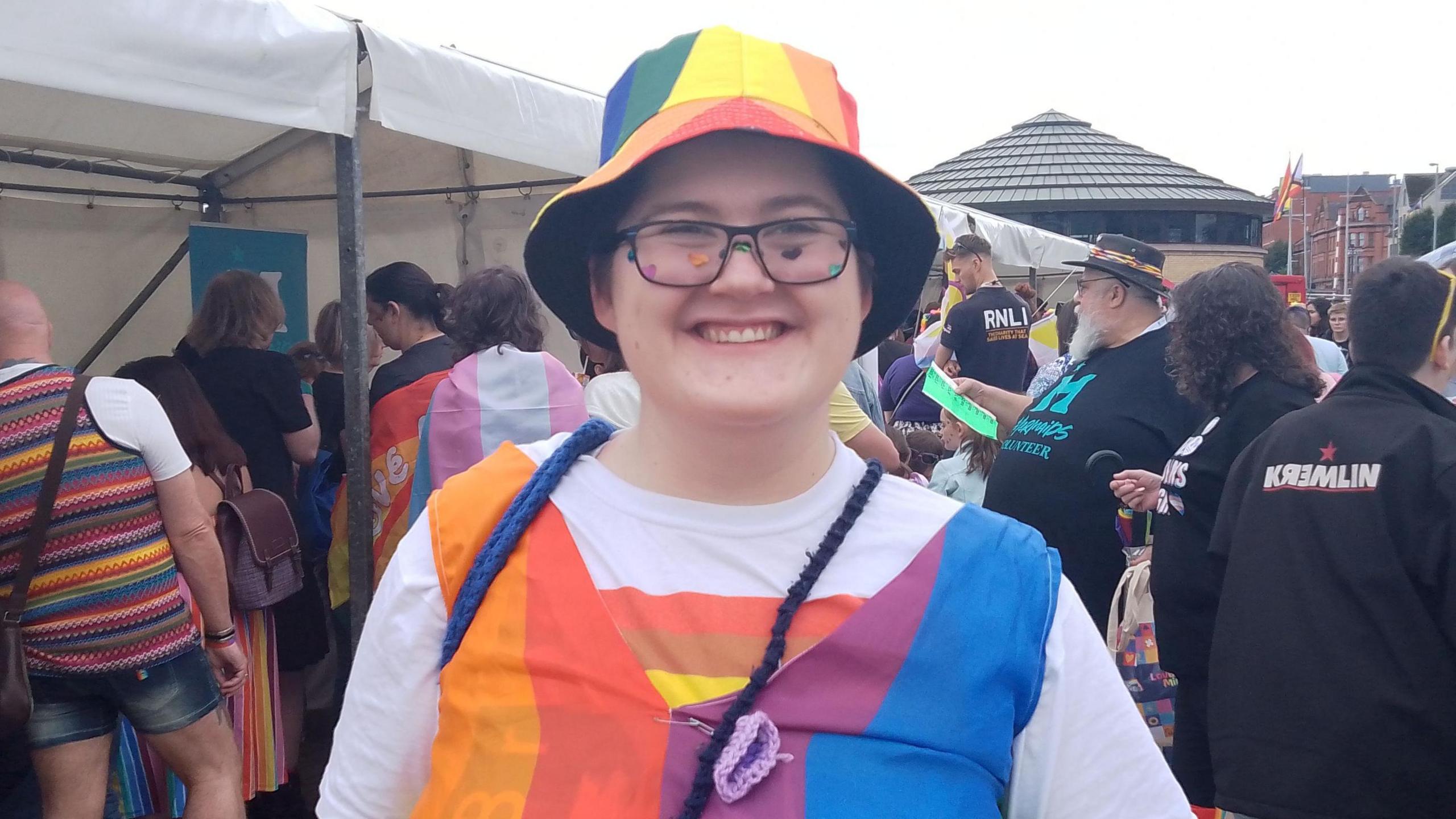 Jackson smiling into the camera wearing a rainbow hat and top. People milling about in background 