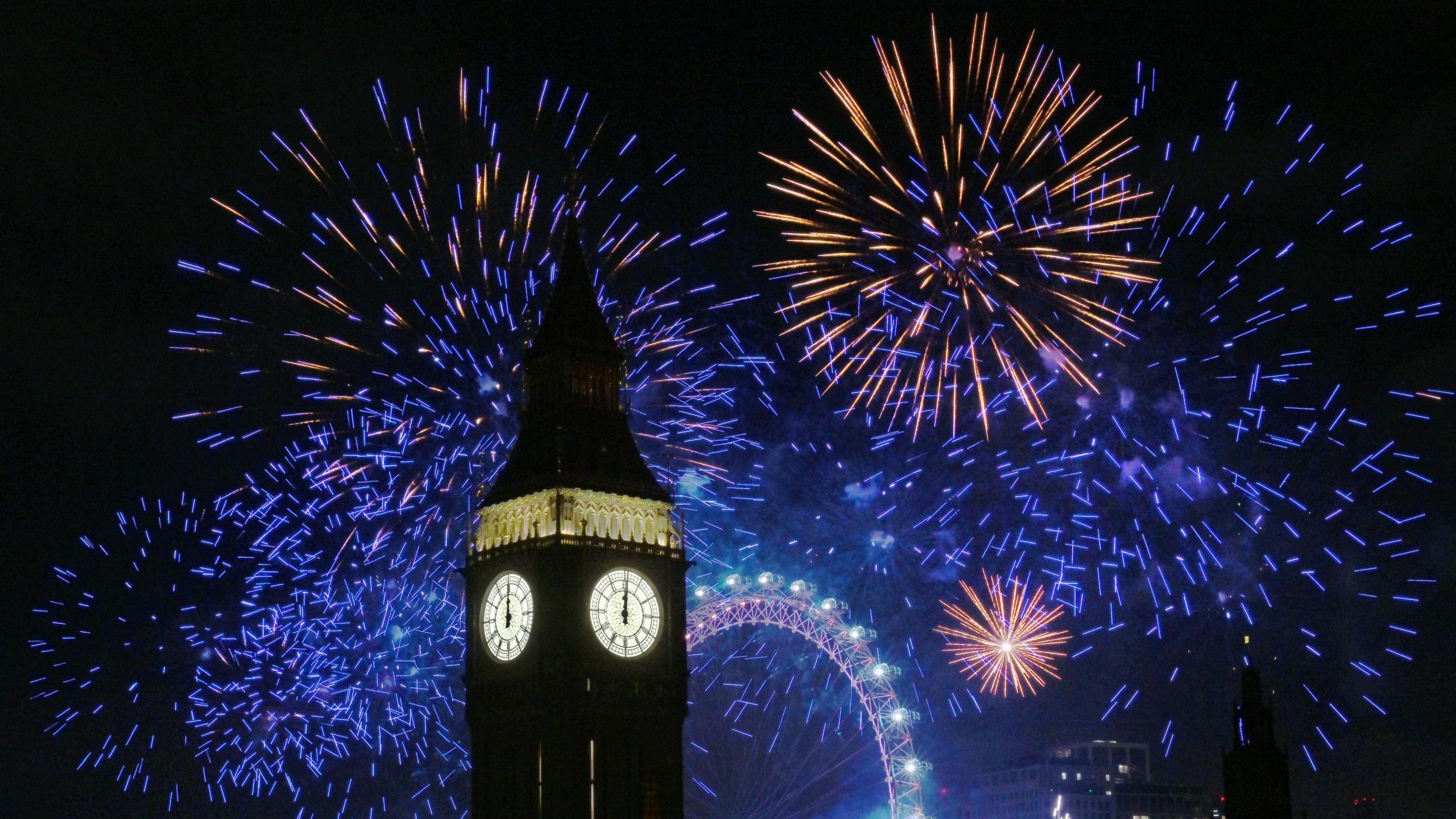 Blue, purple and yellow fireworks exploding against a dark night sky around Big Ben with the London Eye lit up in the background.