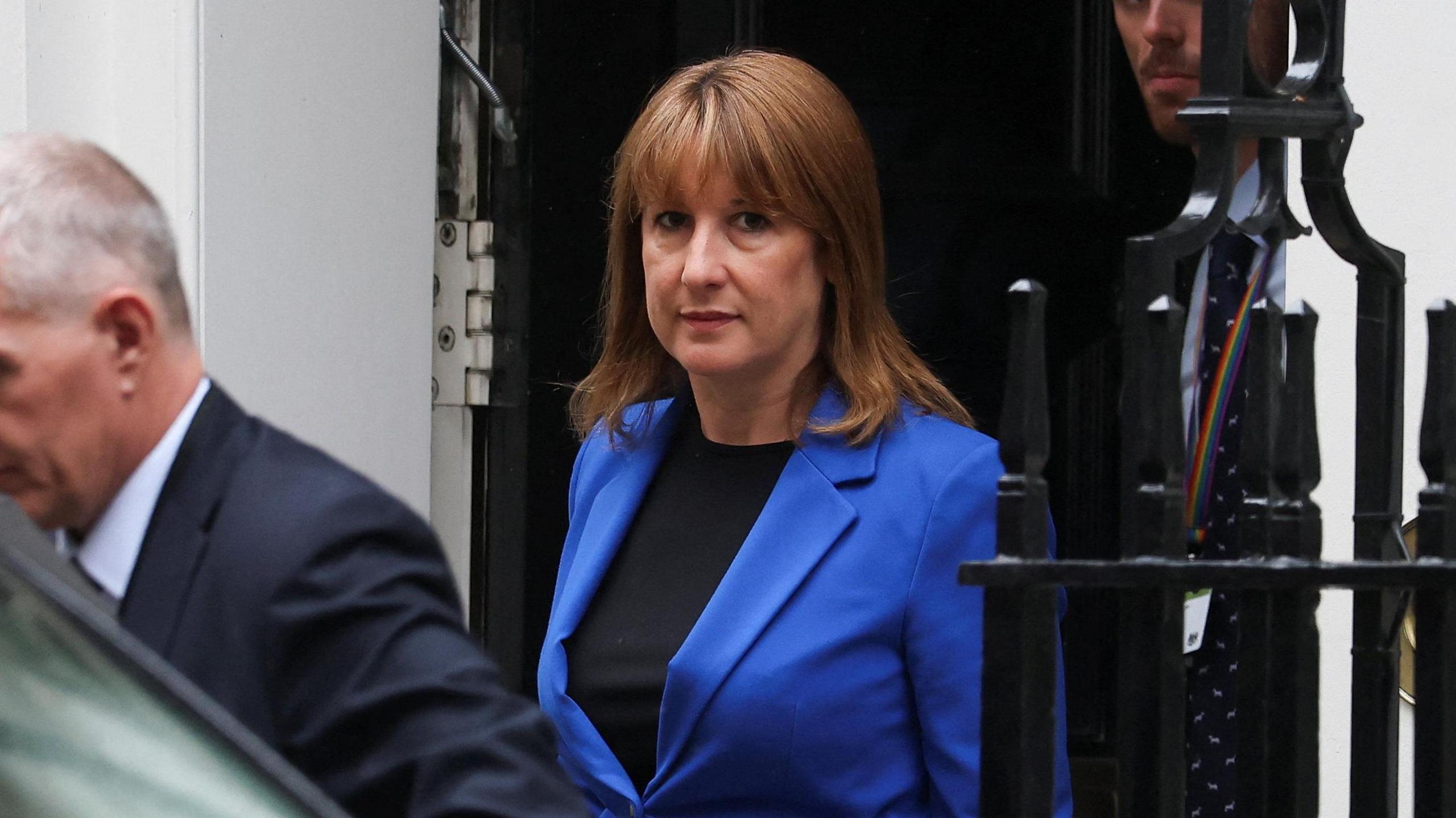 Rachel Reeves, wearing a dark blue suit and black top underneath, exiting 11 Downing Street. There is a male, presumably a security guard, in front of and behind her.