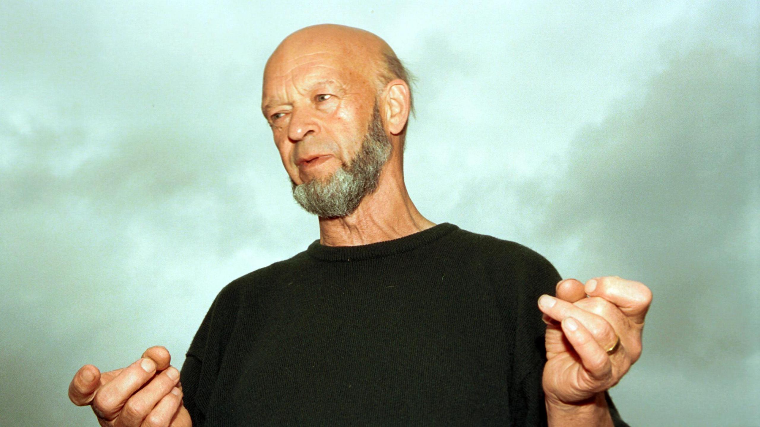 Michael Eavis in a black T-Shirt. He is photographed from slightly below and there are grey clouds behind him.