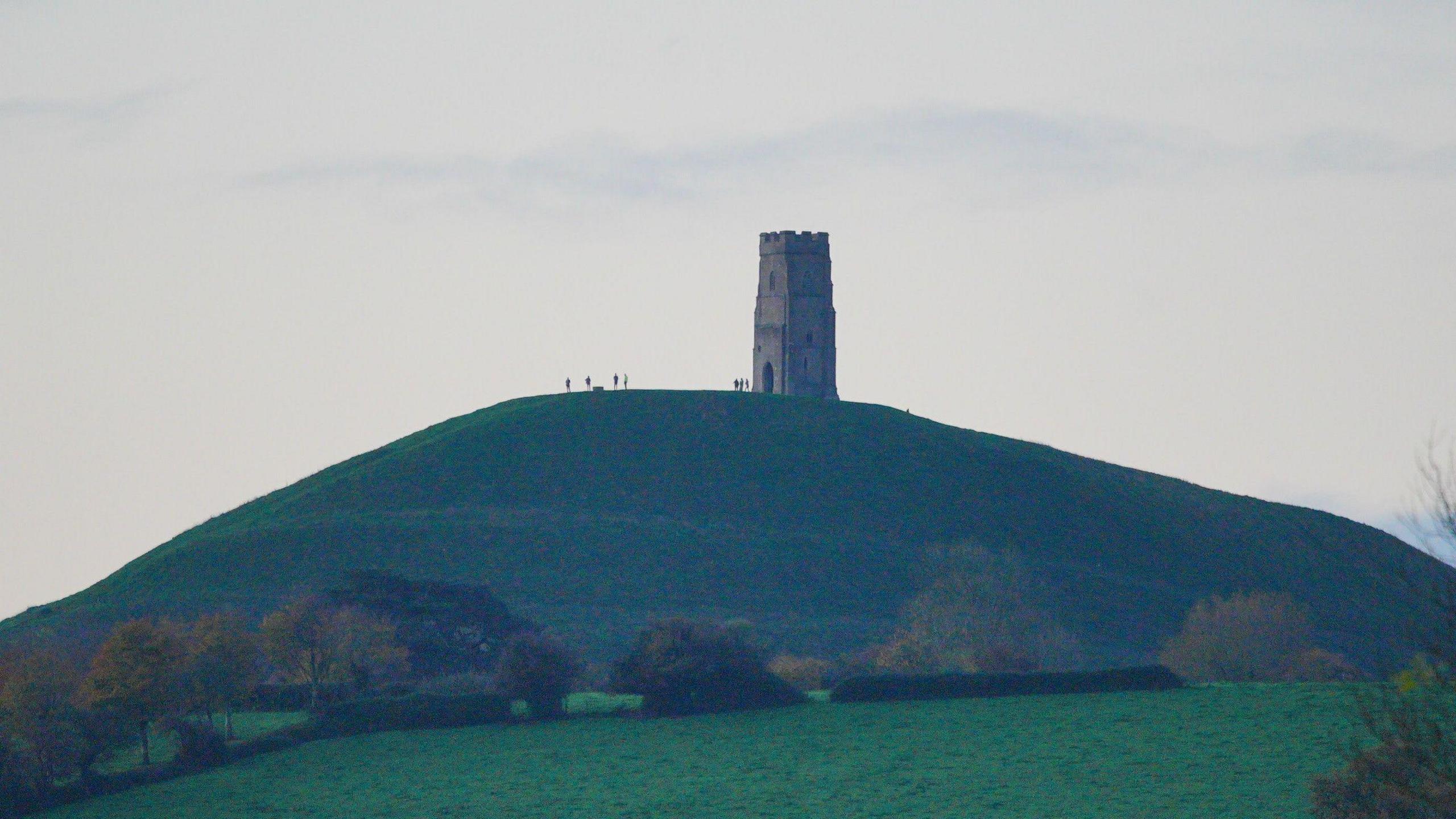 The sun rises over Glastonbury Tor, which is seen in the distance. People's outlines can be seen on the hill.