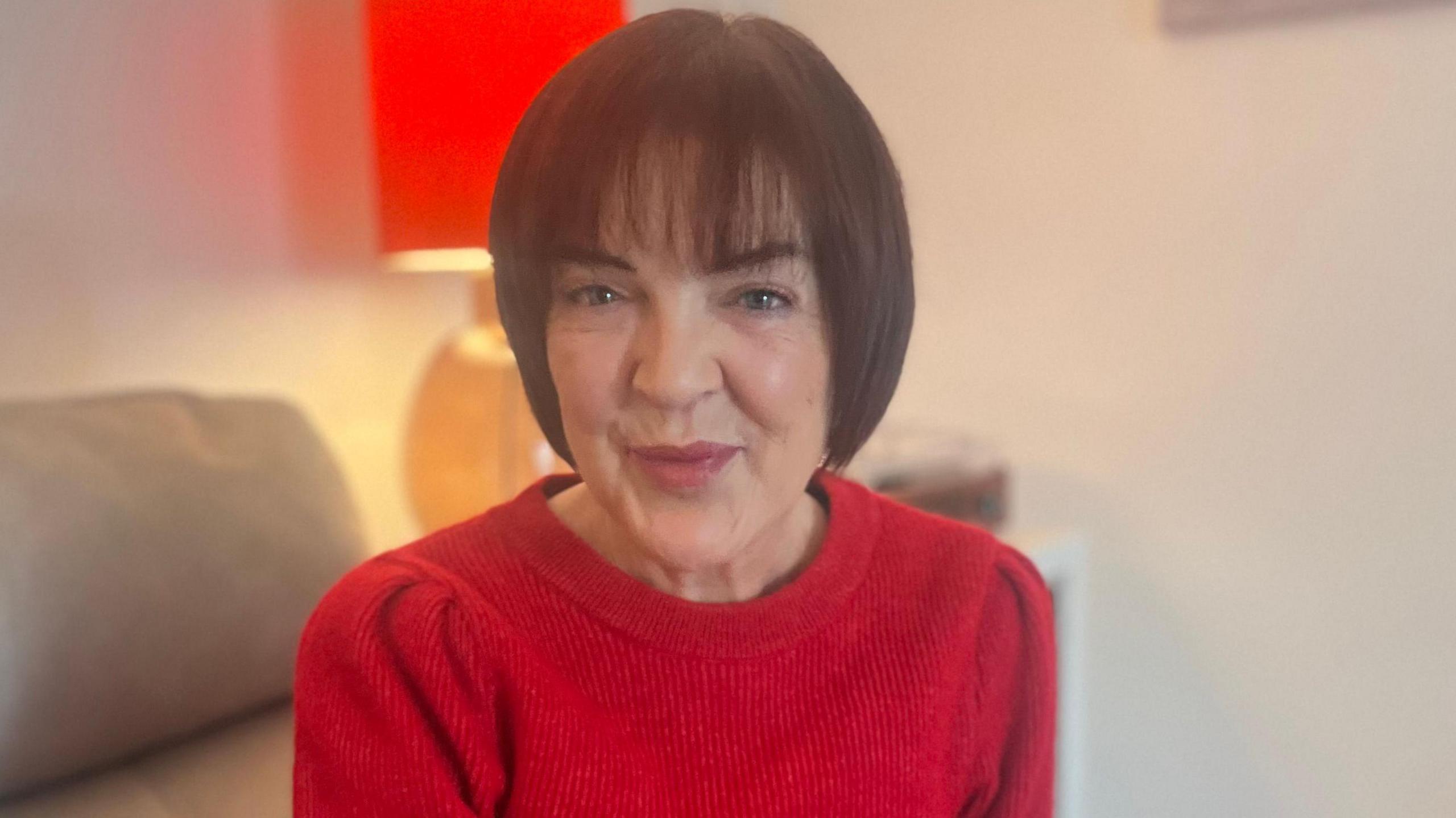 Jennifer has brown hair which is styled in a bob and a fringe. She is sitting on the sofa wearing a red round-neck top. Behind her is a lamp which has a red lampshade. She is wearing makeup.