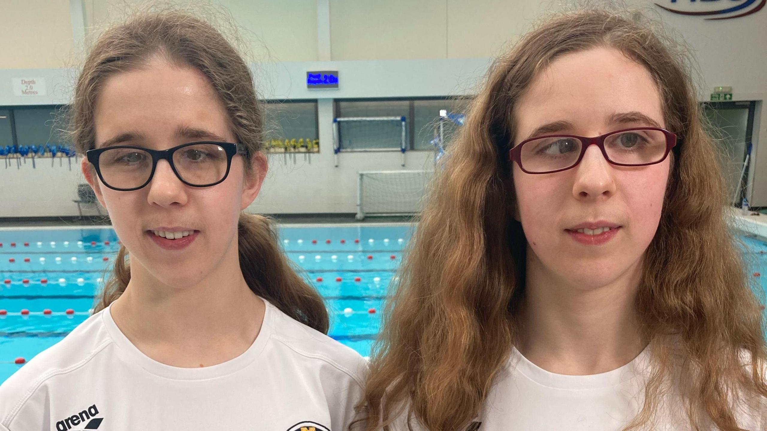 The swimmers are positioned in front of the camera with a pool behind them. They both have long brunette hair - Eliza has her hair in a ponytail, while Scarlett's is down and is curly. They are both wearing glasses and are smiling.