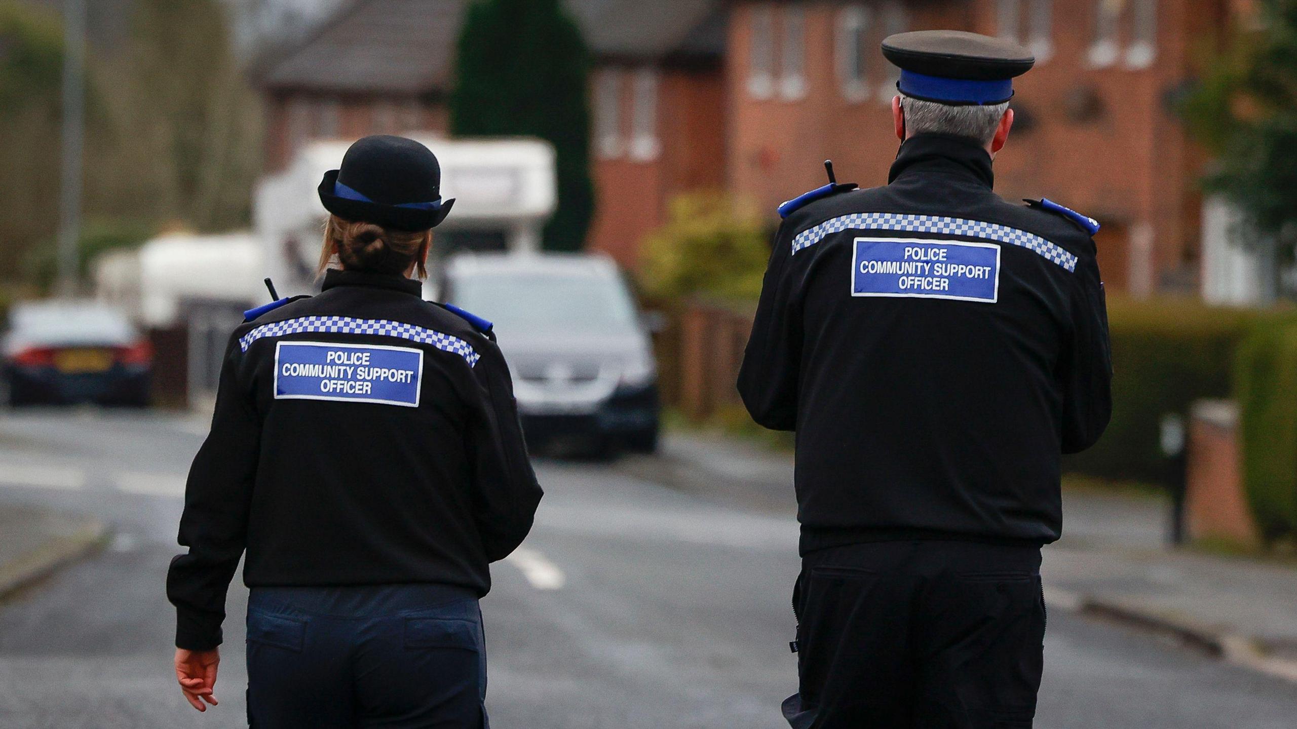 Two police community support officers wearing a black uniform with blue and white chequered detailing on the shoulders, are walking away from the photographer. On the left is a female officer wearing a bowler-style police hat and on the right is a male officer wearing a flat-style police hat. They appear to be walking down the centre of a residential street.