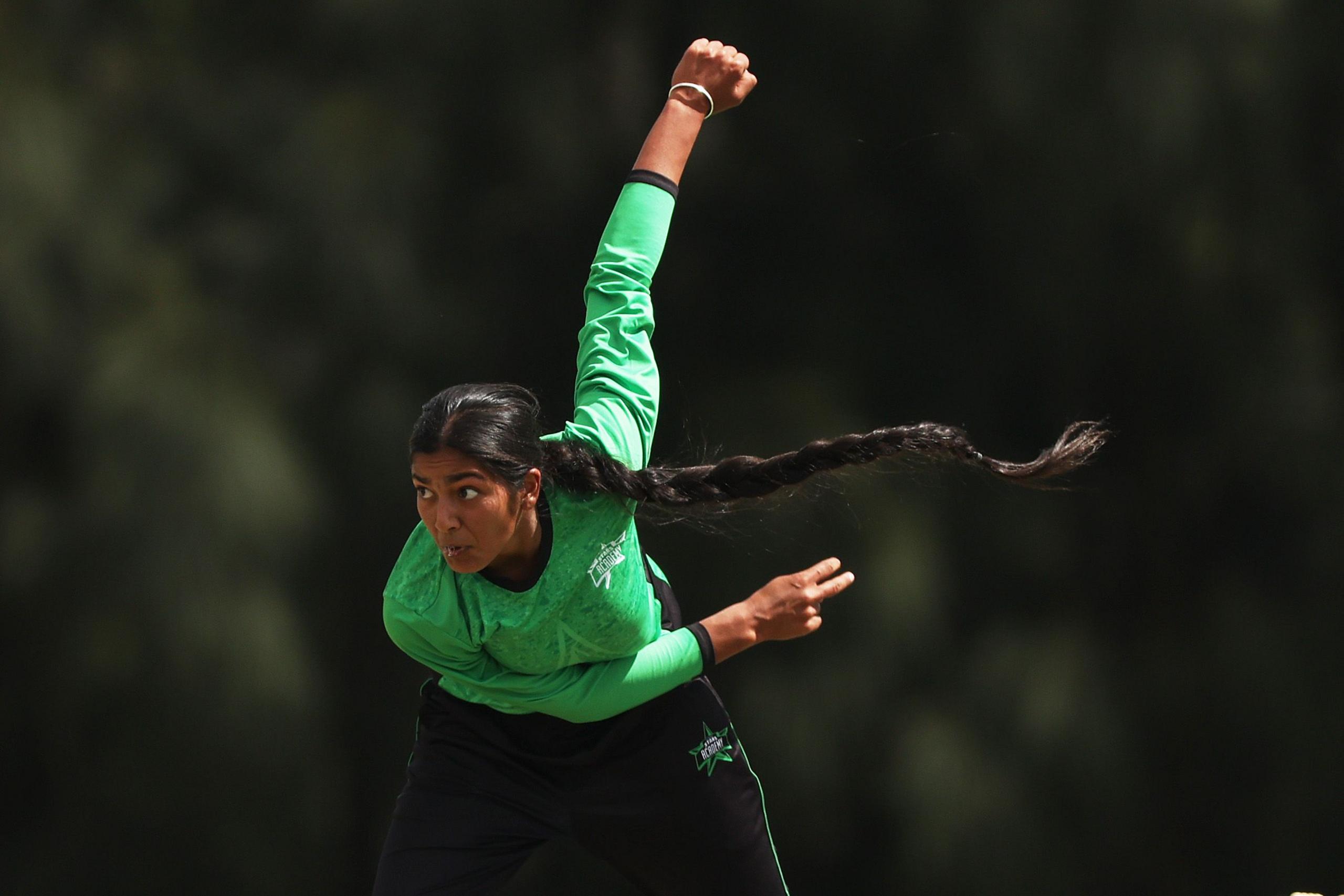 A cricket bowler in a green jersey and black trousers delivers a ball with an overarm action, right arm extended high and left arm bent forward. The player’s long braid swings outward, set against a blurred dark green background.