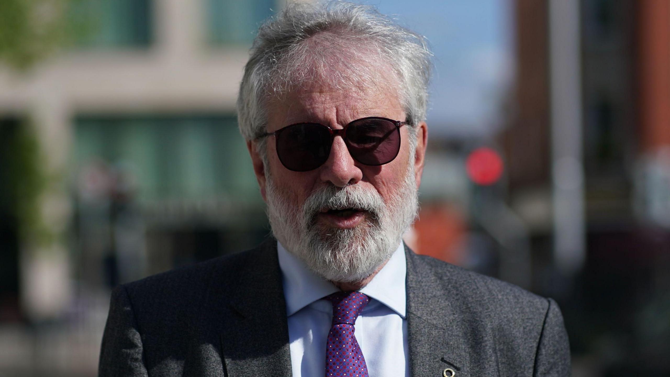 Gerry Adams with grey hair and sungalsses. He is wearing a grey suit, blue shirt and purple tie. He is outside.