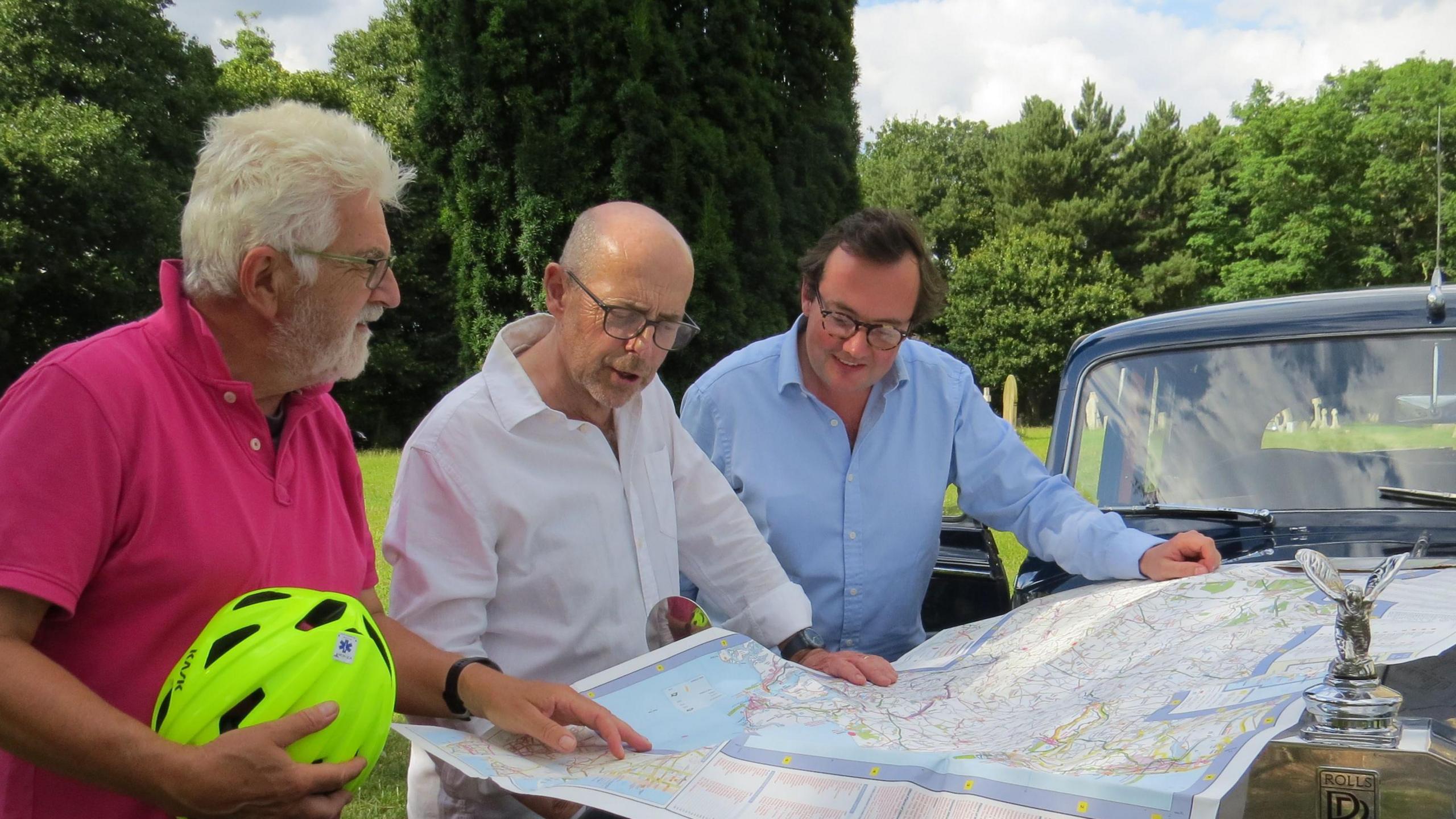 Three men are pictured looking at a large map resting on a car. The man closest to the camera has white hair and is wearing a pink polo top while he holds a florescent green cycle helmet. The man in the middle is bald with a white shirt while the man on the right has dark short hair and is wearing a light blue shirt.