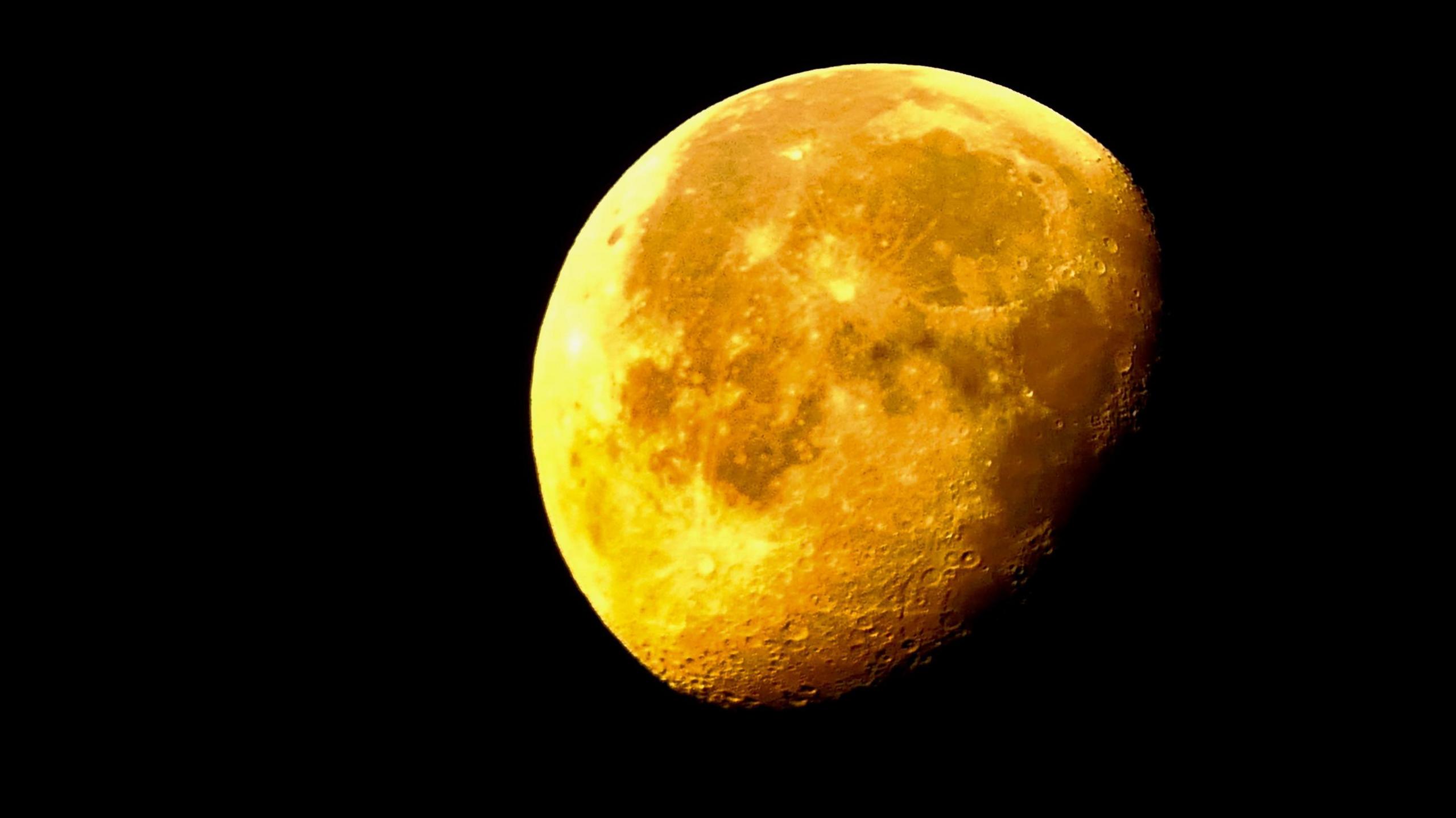 A close up of the moon which has different shades of yellow and orange. A quarter of the moon, on the right, is dark from a shadow. 